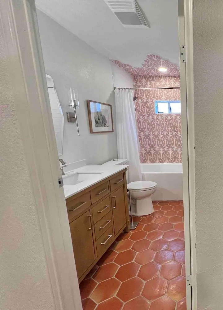 Bathroom featuring hexagonal terracotta-toned floor tiles, a light wood-finish vanity with dual sinks and white countertops, a built-in tub with pink patterned wall tile, and wall-mounted sconce lighting