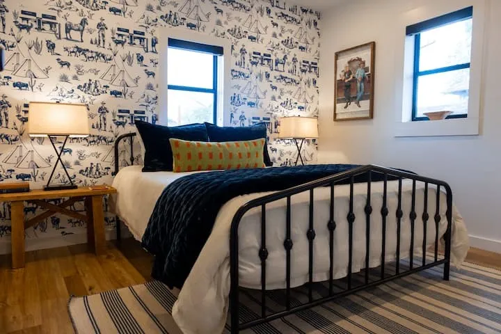 Spacious room featuring wood-finish flooring, two windows with black roller shades, and a feature wall with patterned wallpaper