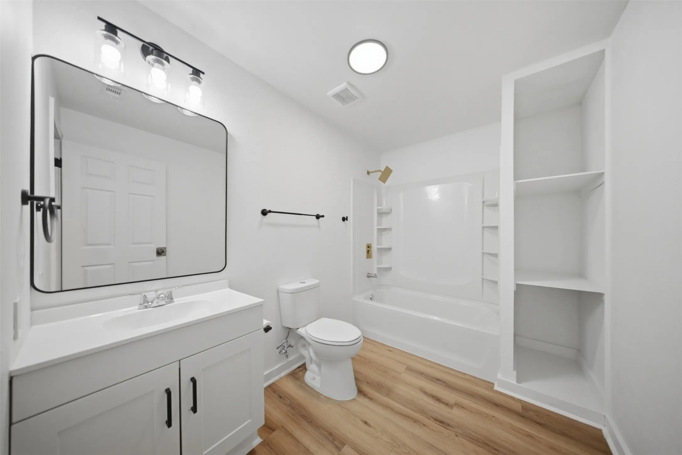 Bathroom featuring wood-finish flooring, a white vanity with cabinet storage, a rectangular framed mirror, a three-light vanity fixture, and a built-in shower-tub combination with gold-tone fixtures