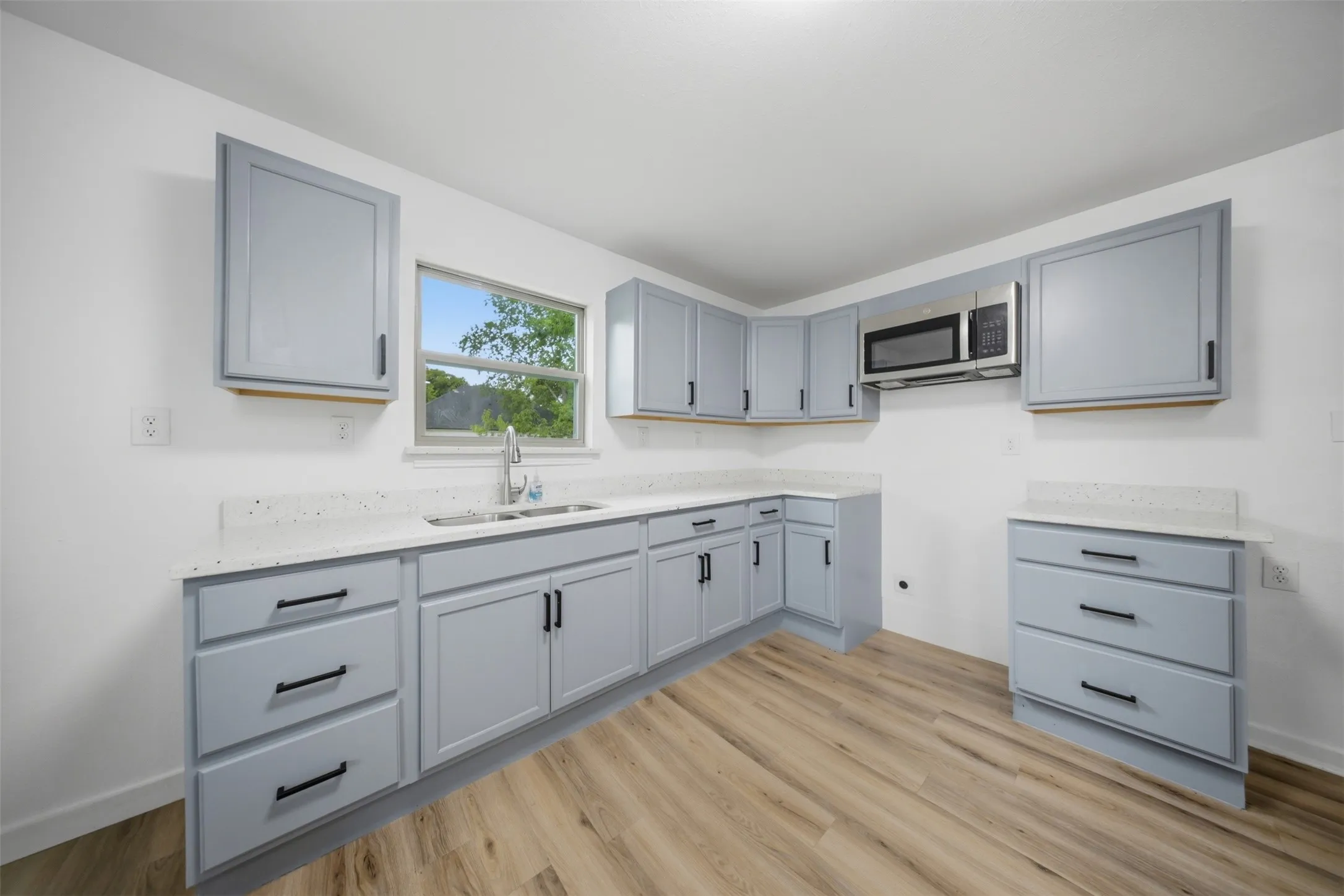 Kitchen featuring light blue cabinetry, white countertops, stainless steel microwave, double basin sink, and wood-finish flooring