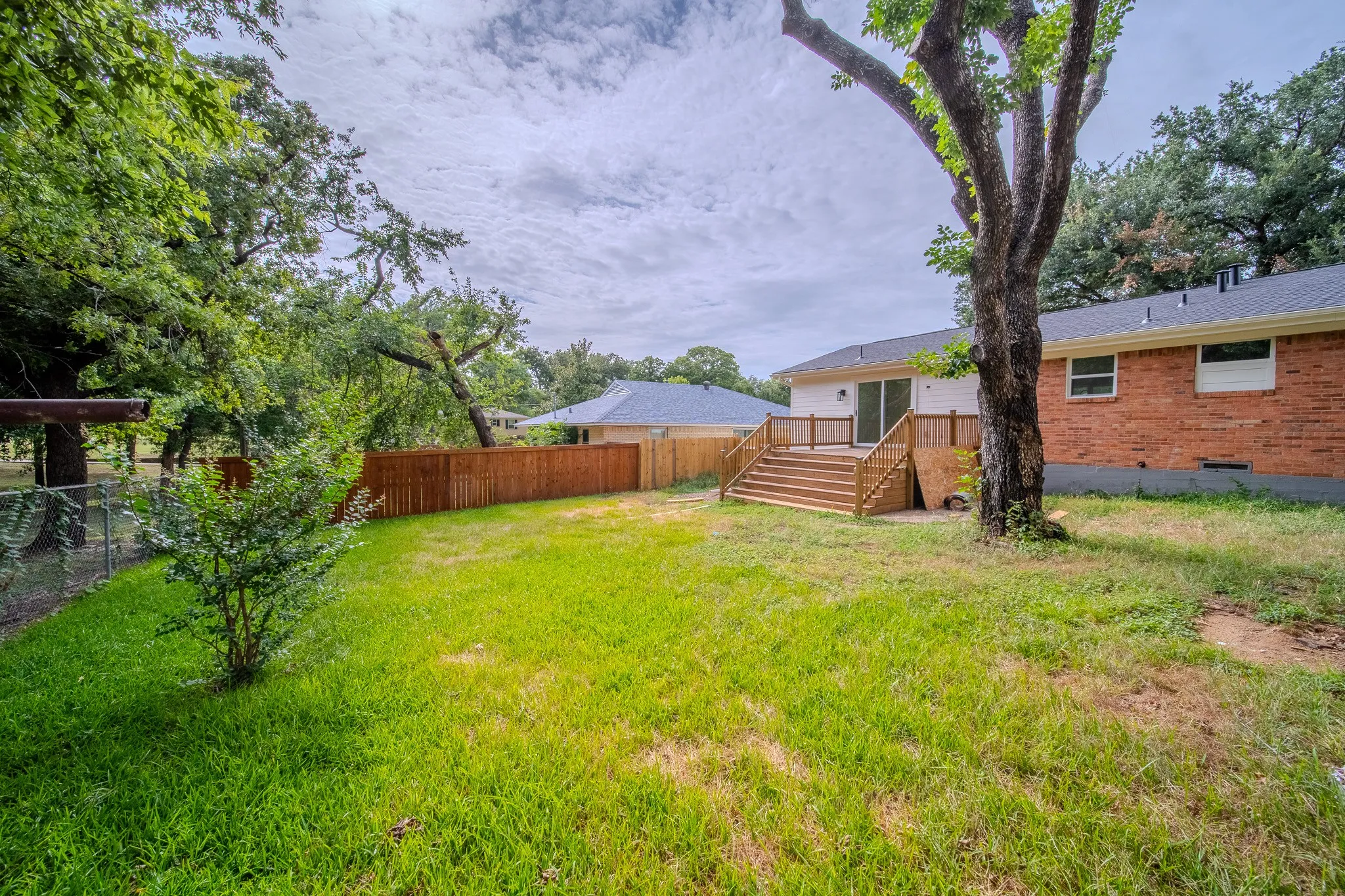Spacious backyard with a wood privacy fence, chain-link fence, mature trees, and a multi-level wood deck featuring white railings and a sliding glass door