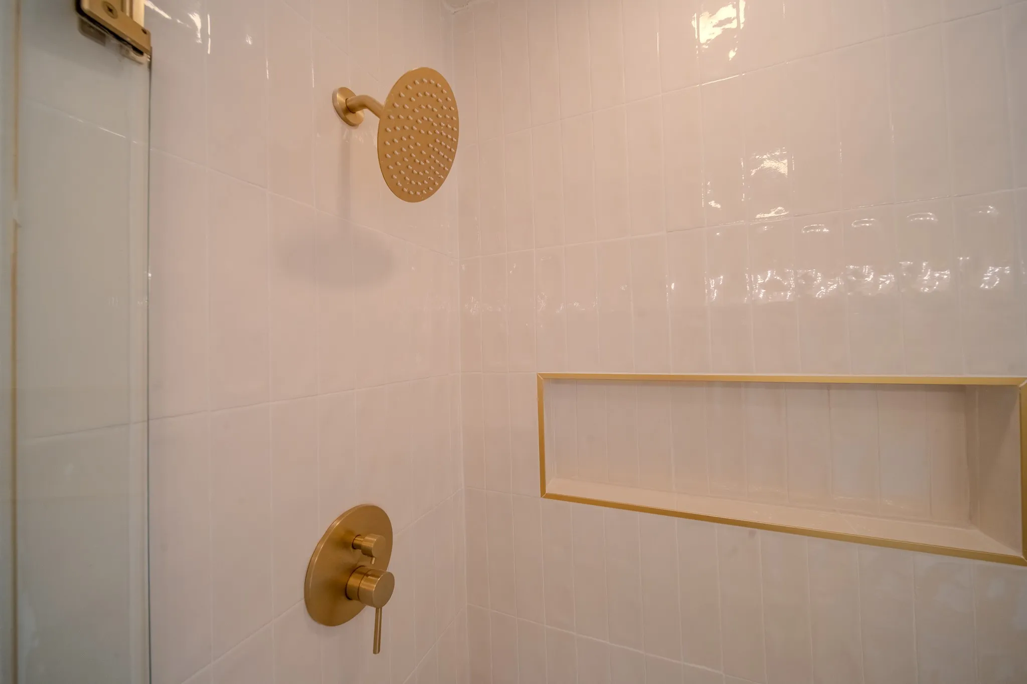 Shower enclosure featuring white subway tile, a gold-finish rainfall showerhead, coordinating gold-finish controls, a glass partition, and an integrated recessed niche with gold trim