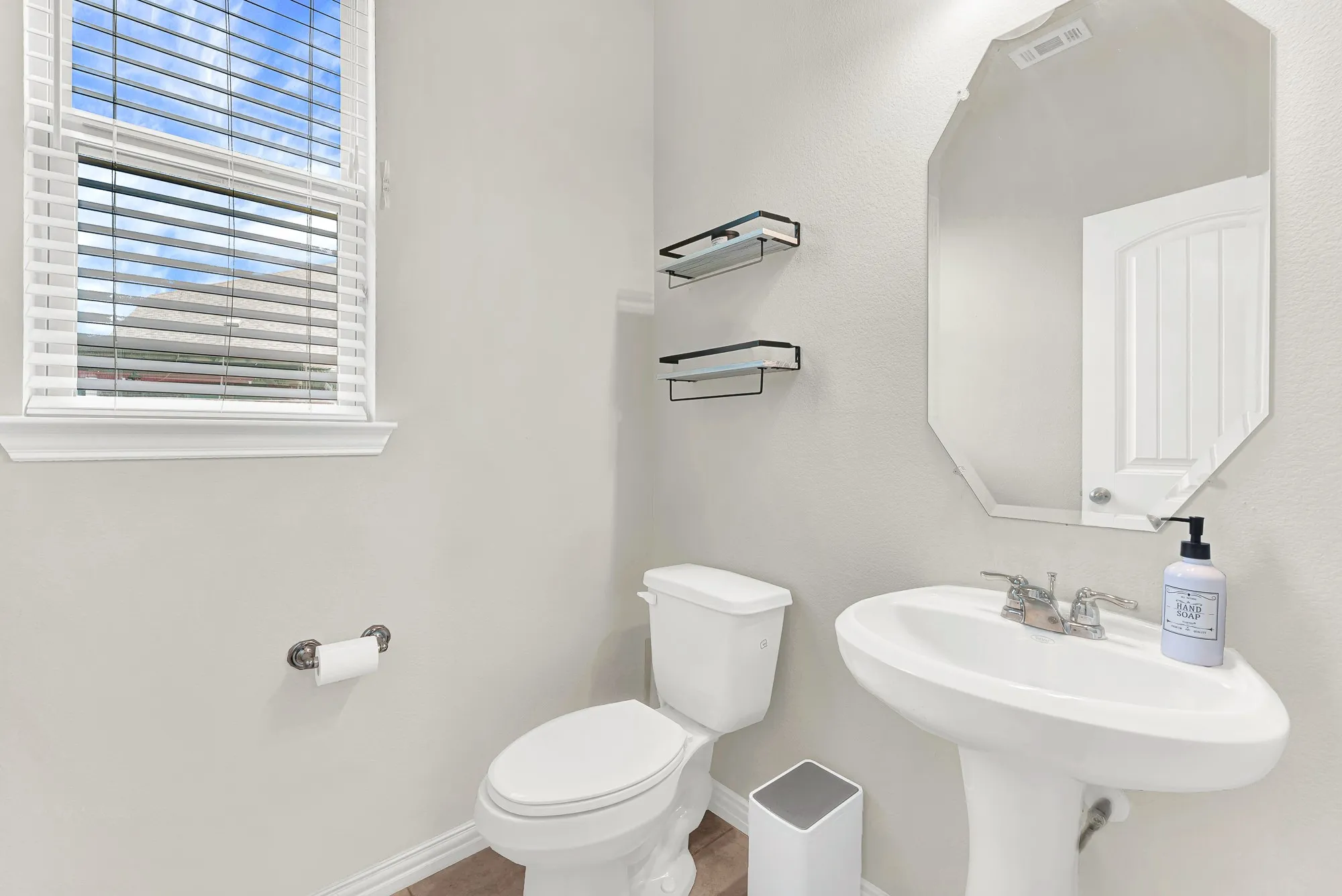 Pedestal sink with chrome fixtures, an octagonal wall mirror, and a window with blinds