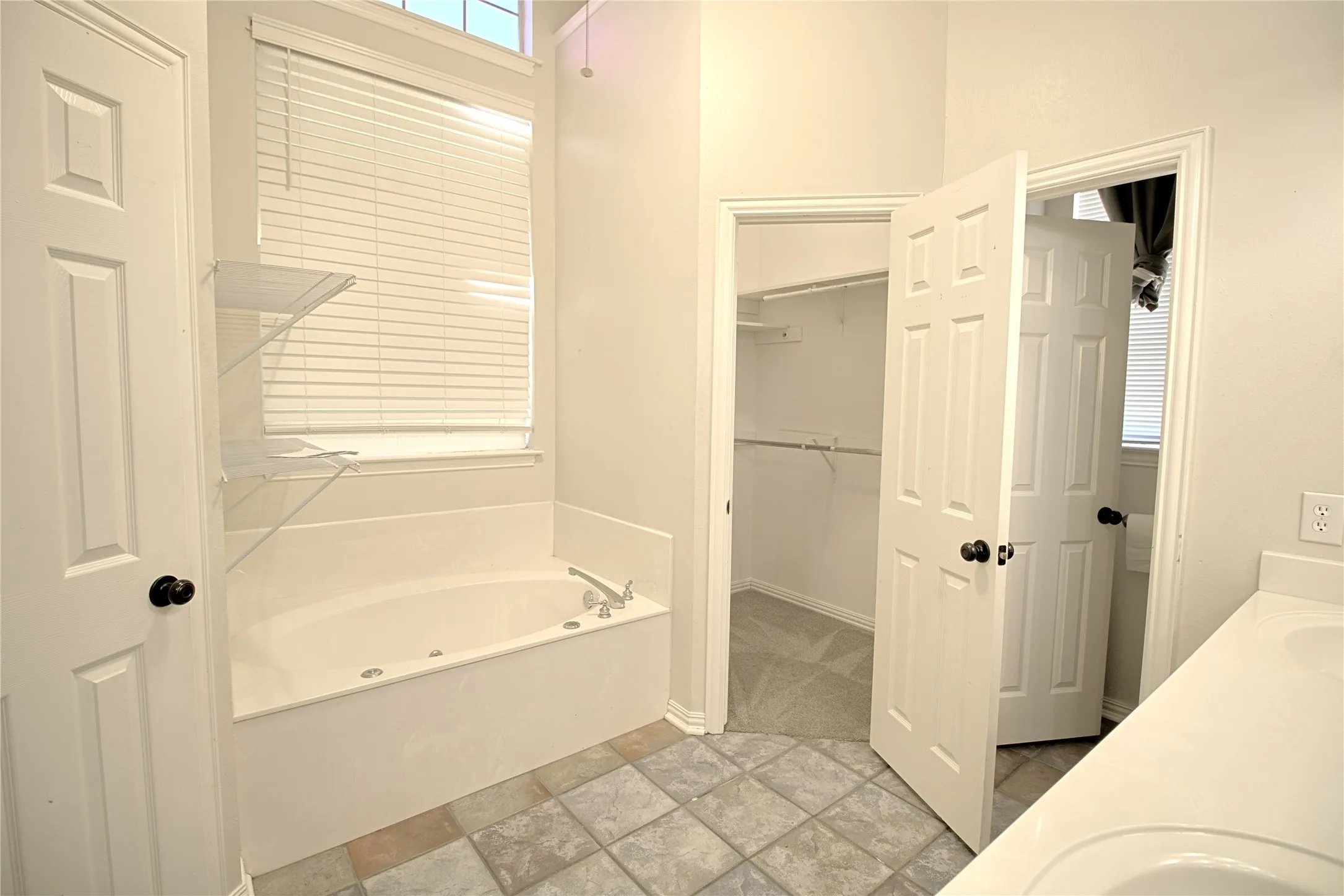 Soaking tub with jetted features, white window blinds, and clear acrylic shelving