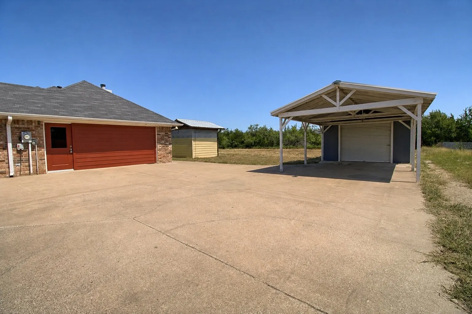 Expansive concrete driveway servicing a two-car garage with a red door and horizontal siding, a detached carport, and an additional storage building