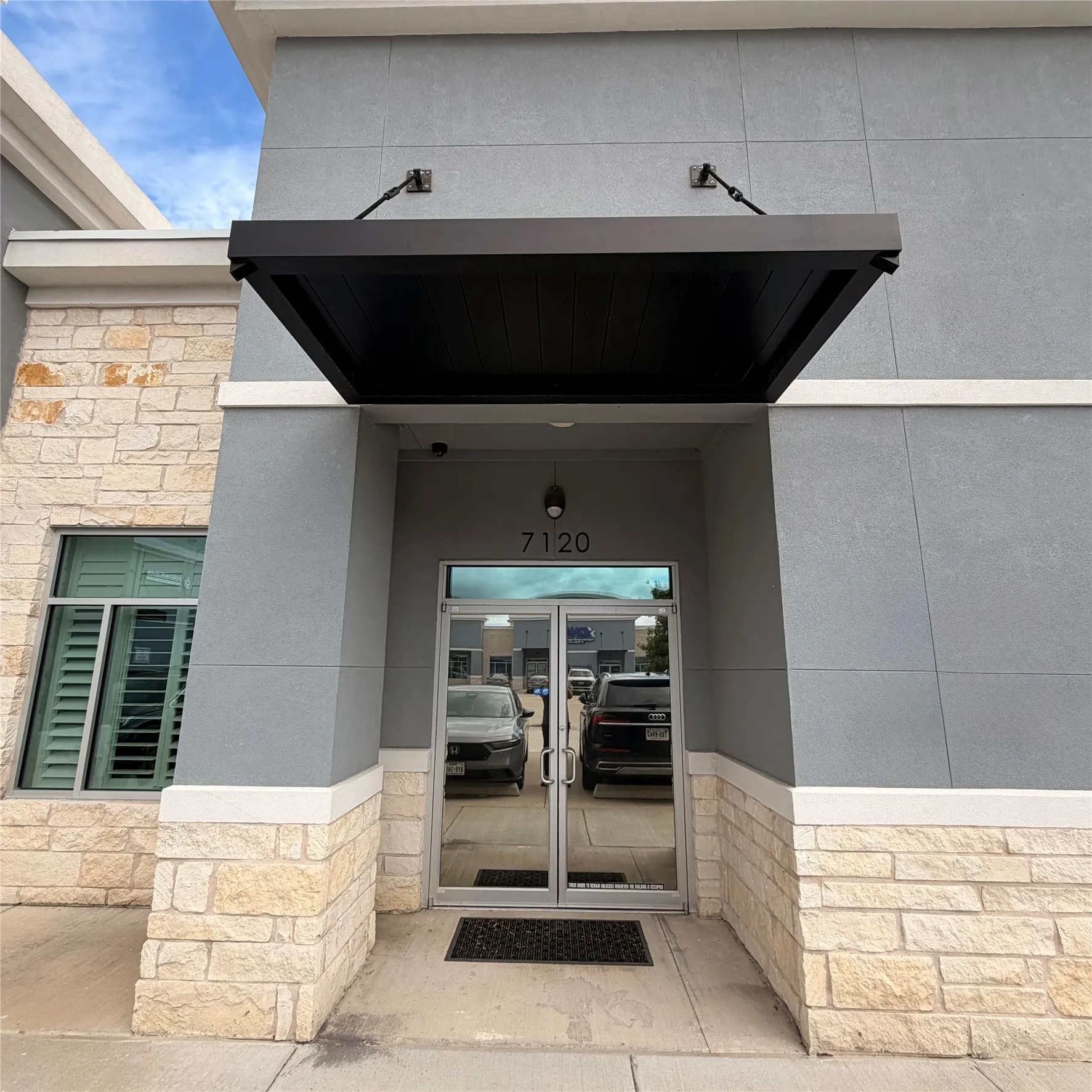 Modern commercial entrance featuring a black overhead awning, prominent building numbers, a light gray stucco facade, and a stone veneer base