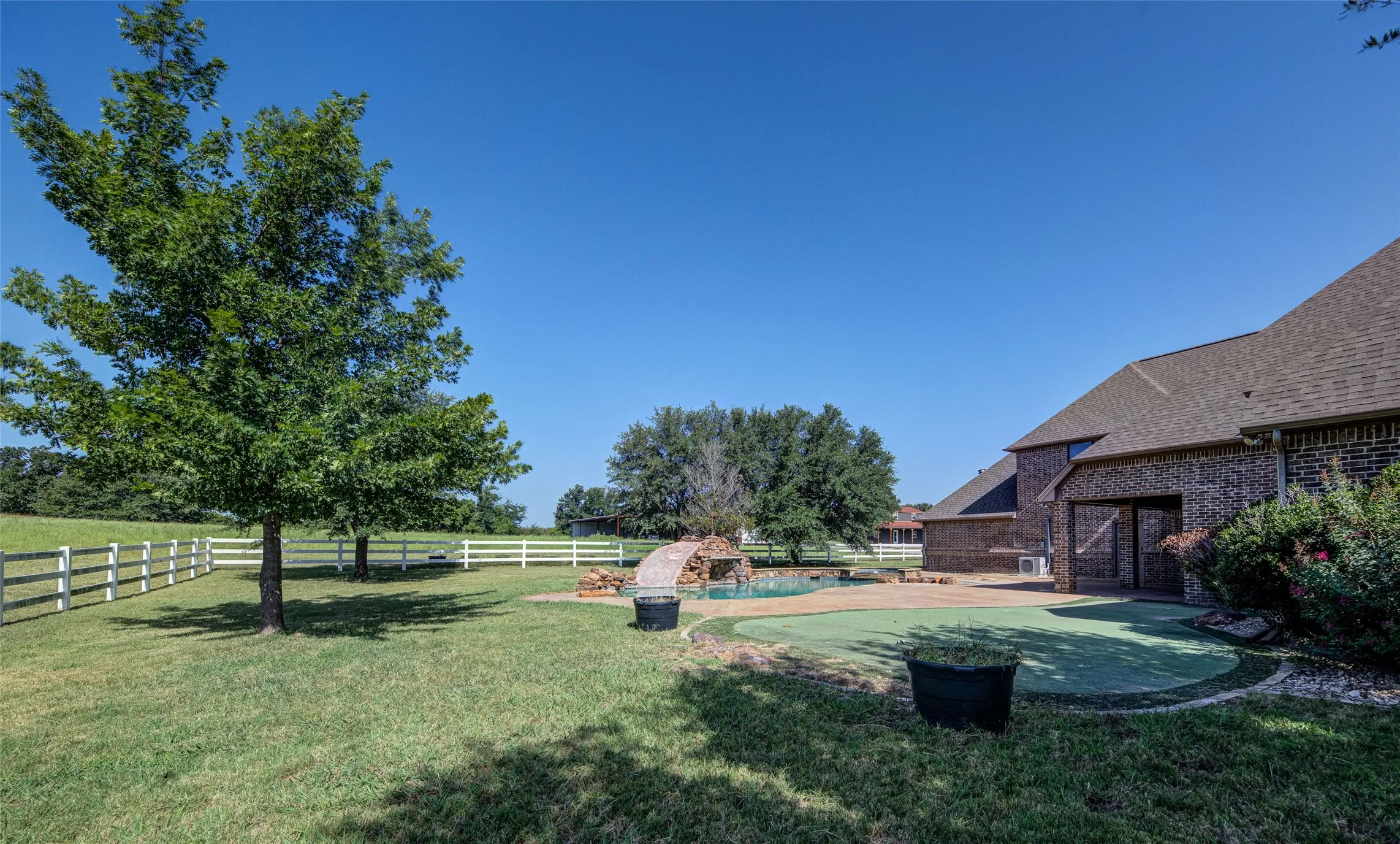 Expansive backyard featuring a manicured lawn, white rail fencing, a swimming pool with a rock waterfall feature, a paved patio, and a brick exterior home