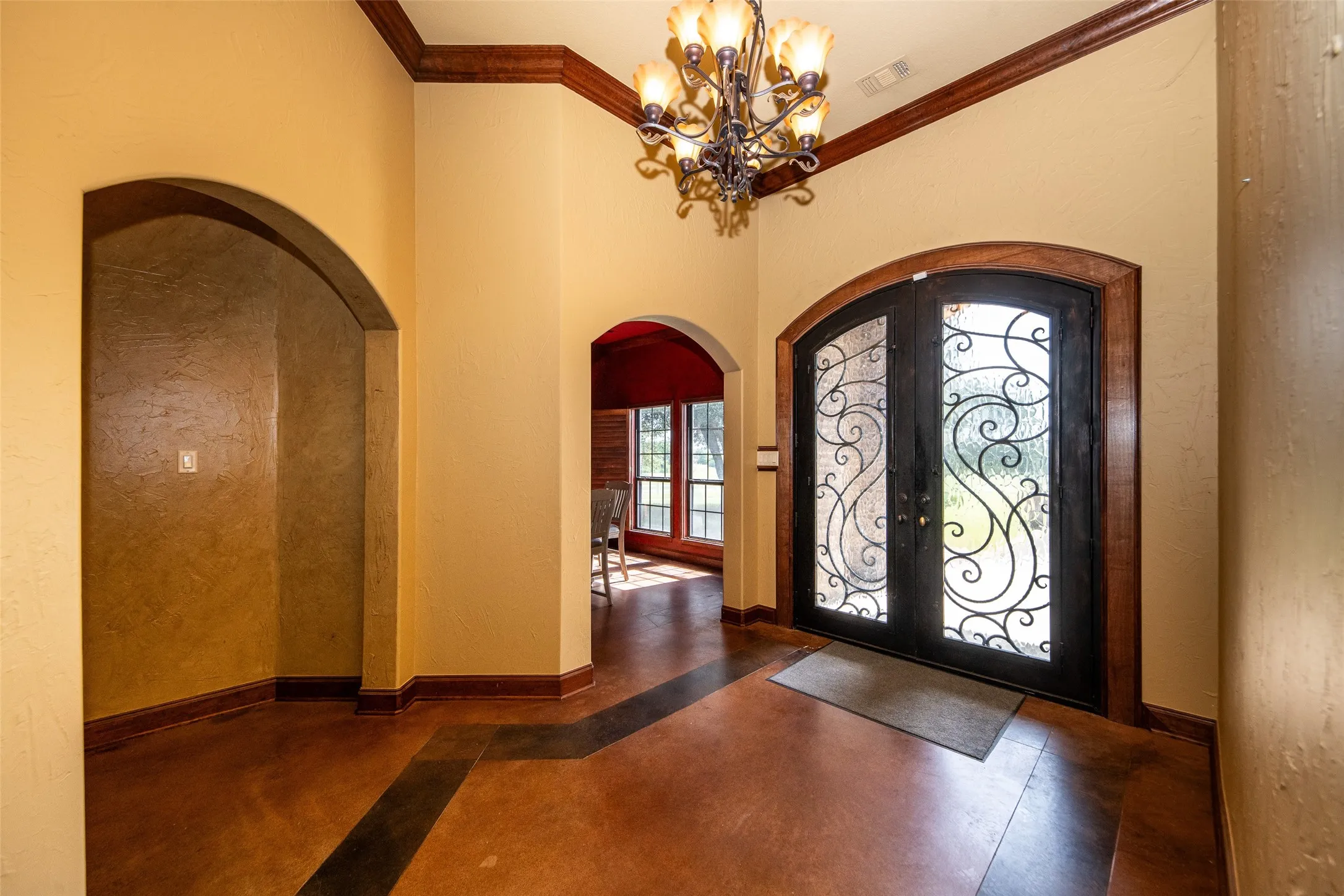 Entryway featuring ornate iron and glass double doors, wood-stained trim, arched doorways, a decorative chandelier, and stained concrete flooring with a dark accent pattern
