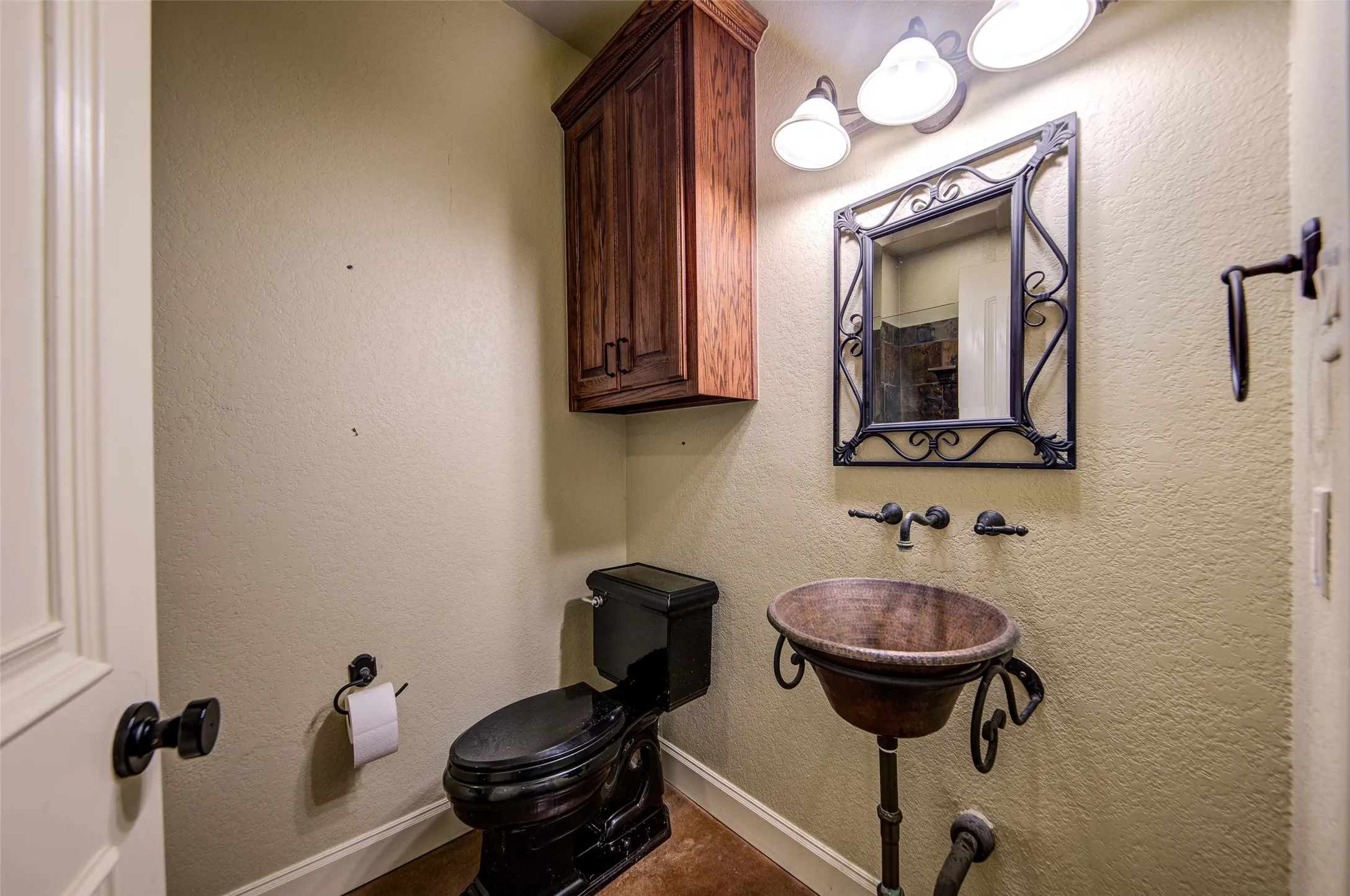 Powder room featuring a unique hammered metal vessel sink with an ornate stand, a black toilet, and a dark wood-finish wall cabinet