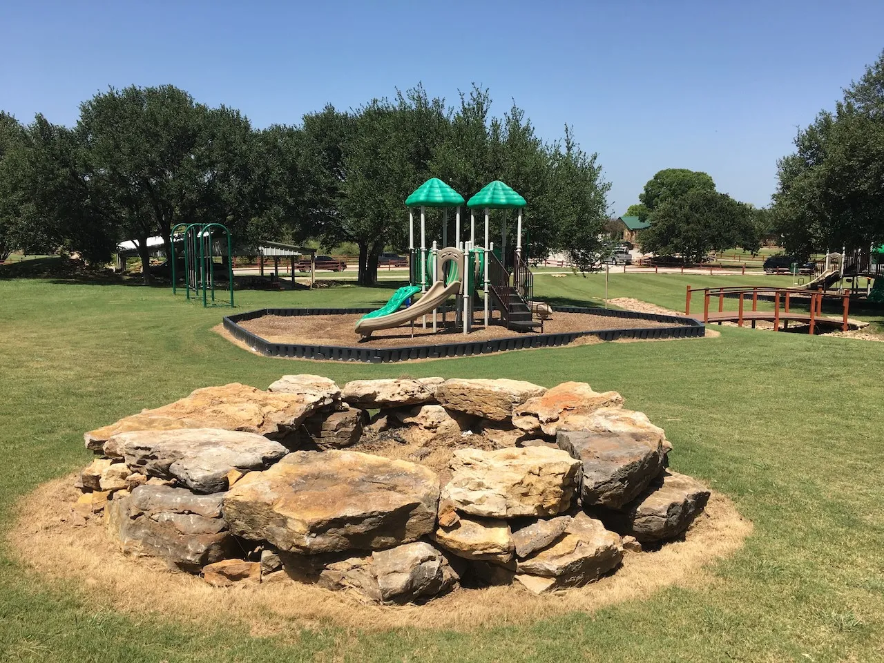 Expansive green space featuring a rock-lined landscape element, a multi-component play structure with slides and climbing features, and a small wooden pedestrian bridge