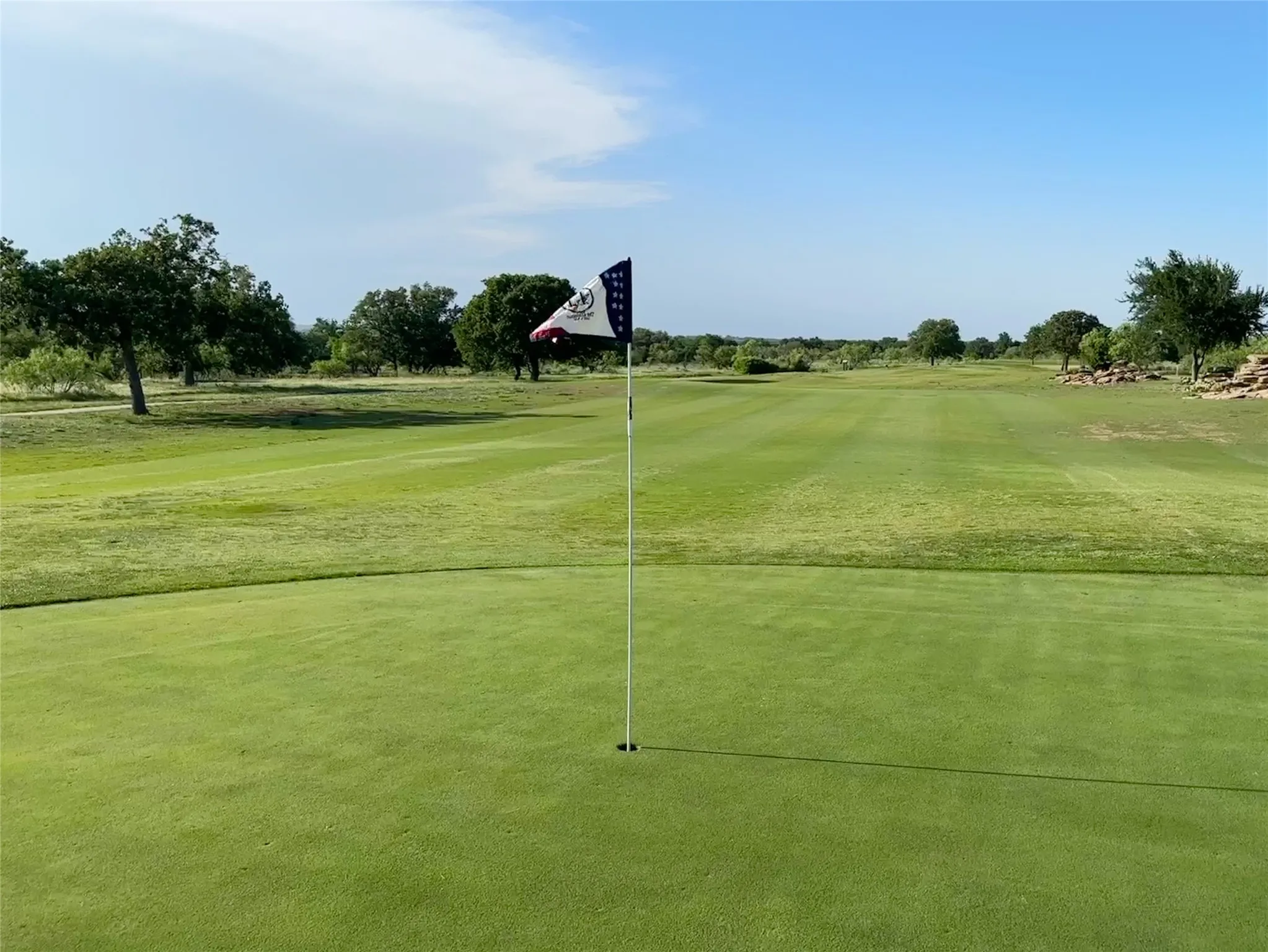 Manicured golf green with flagstick, surrounded by a fairway, mature trees, and natural rock formations