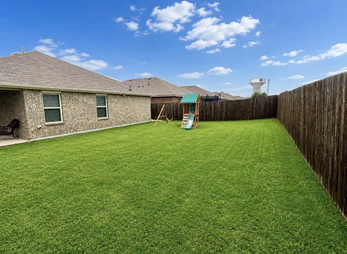 Expansive backyard featuring a manicured lawn, full privacy fencing, and a brick exterior with a covered patio
