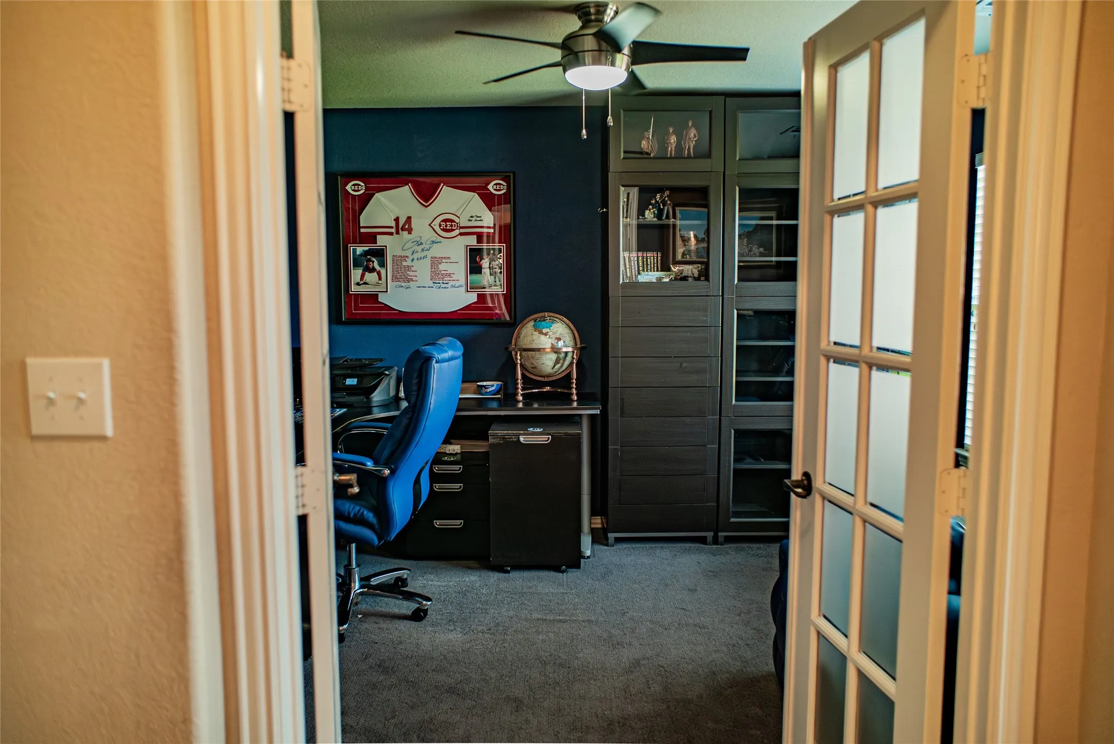 Double French doors opening to an interior room with a ceiling fan, wall-to-wall carpeting, and built-in cabinetry with glass-fronted sections