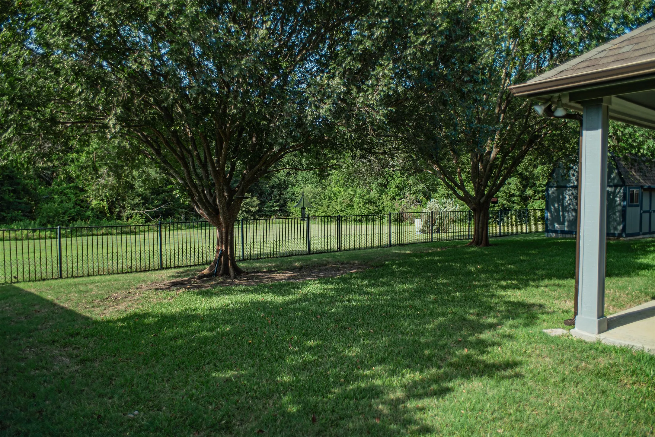 Expansive grassy yard featuring mature trees, a black metal fence, and a covered patio area with a light fixture