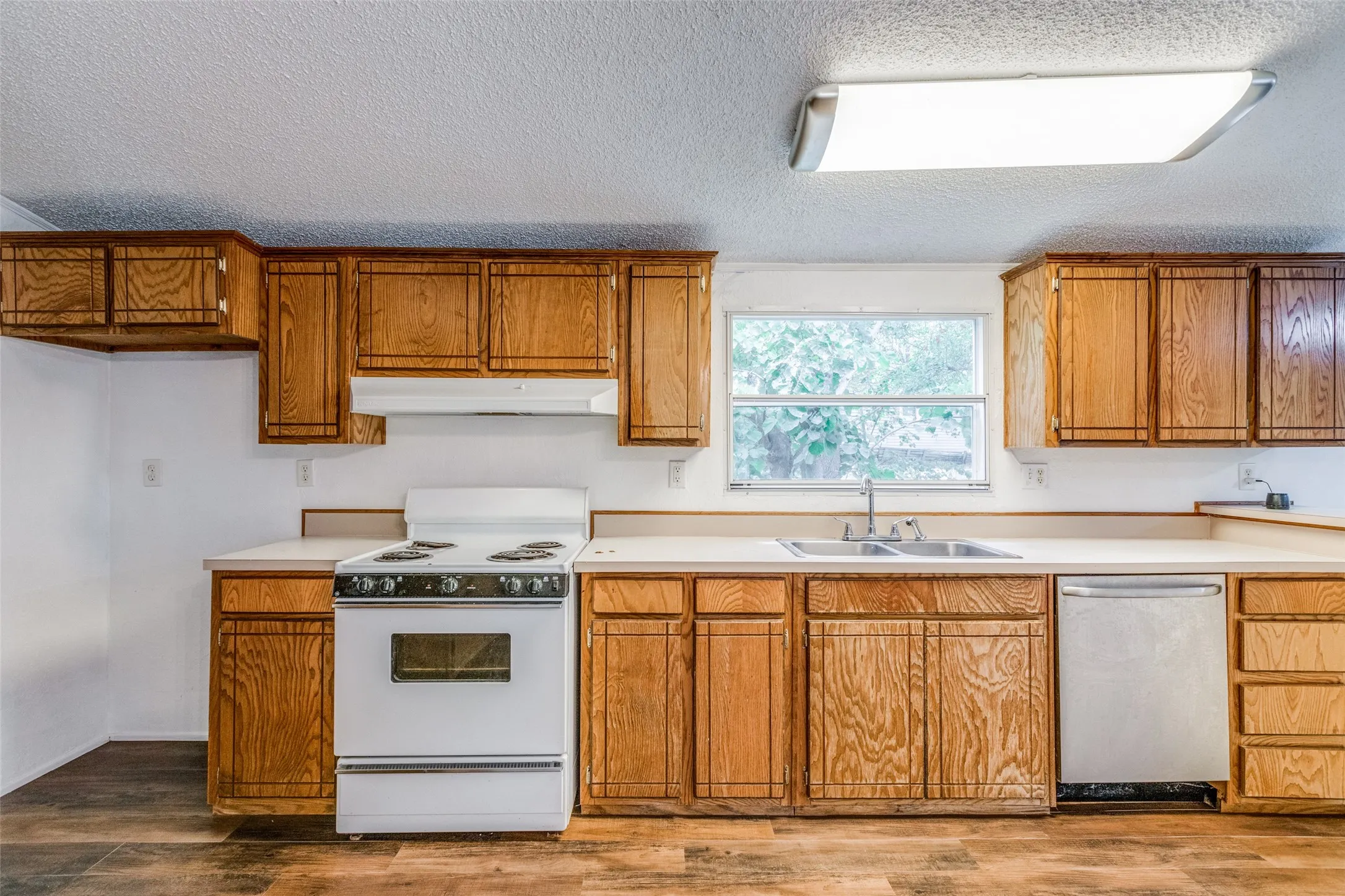 Kitchen with space for a refrigerator, space for a range, stainless steel dishwasher, wood-type flooring, and a textured ceiling.