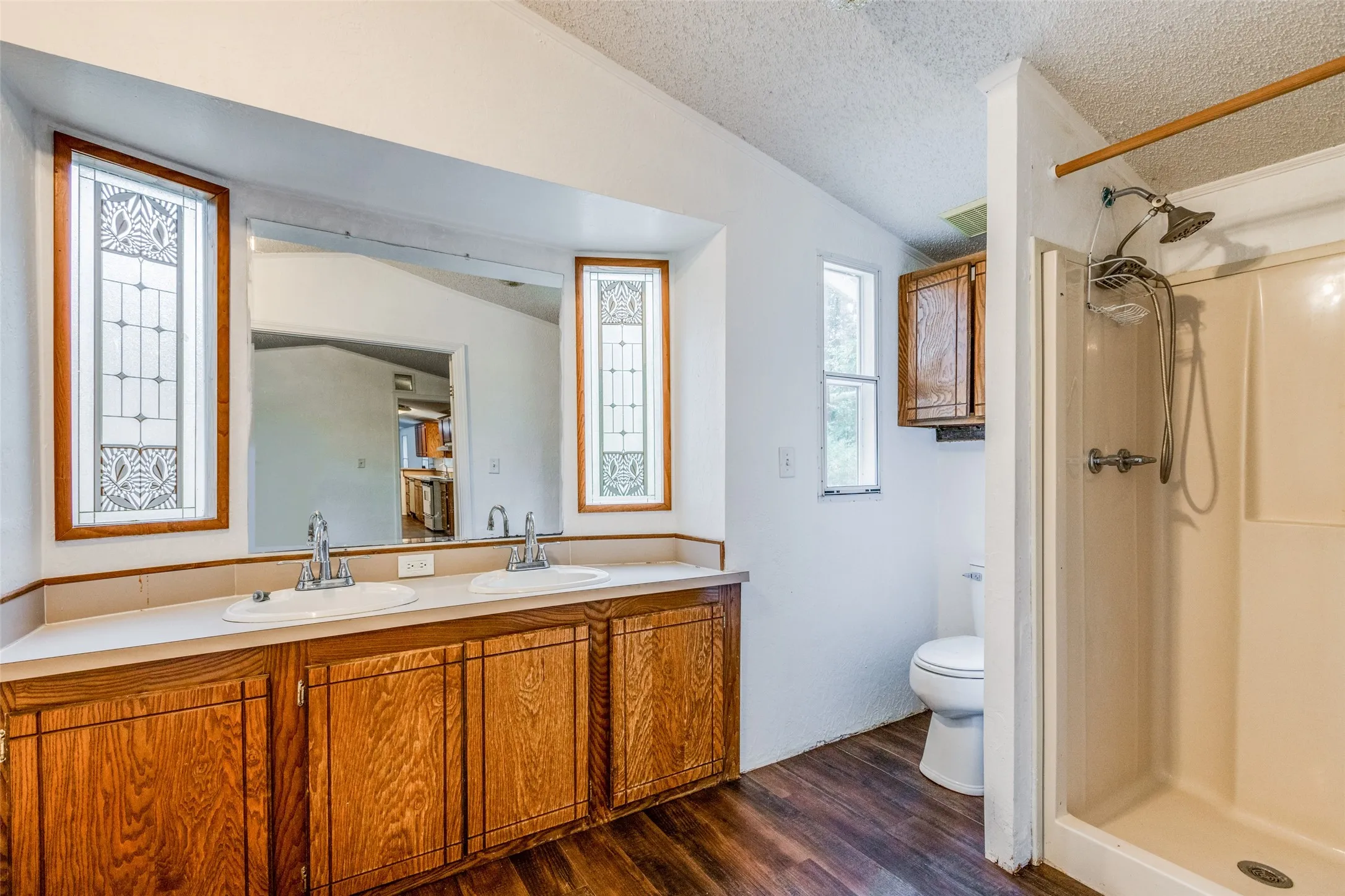 Primary bathroom with hardwood/ wood-style flooring, double sink vanity, walk in shower, vaulted ceiling, and a textured ceiling.