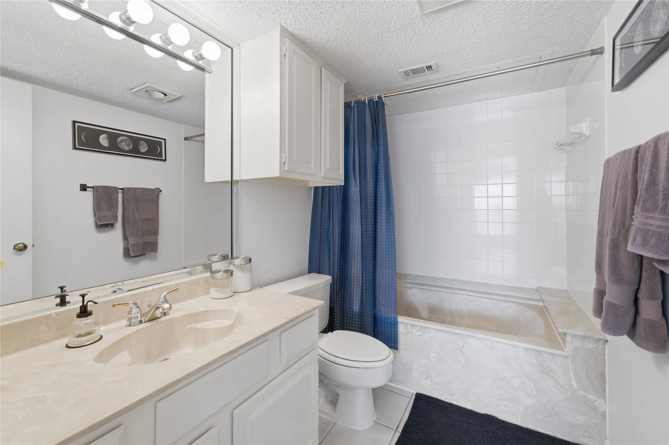 Bathroom featuring a single vanity with a light-toned countertop, white cabinetry, a built-in bathtub with white tile surround, and an overhead vanity light bar