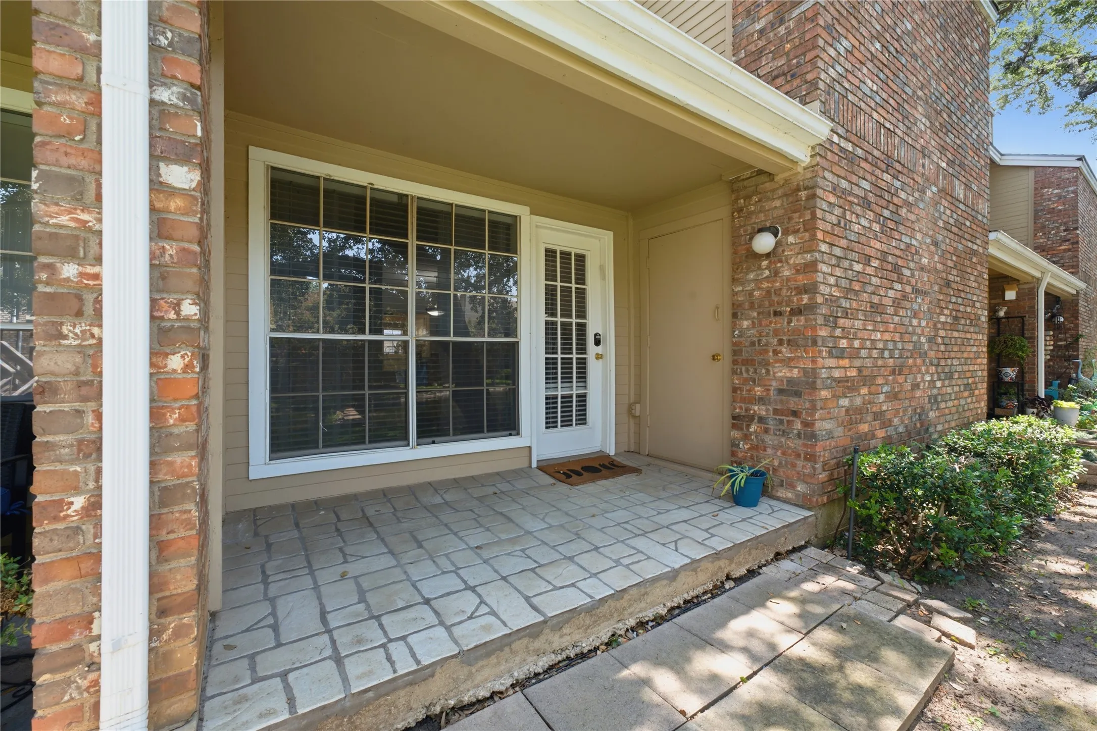 Brick exterior featuring a covered entry, decorative paver patio, full-light entry door, and large window with grid panes