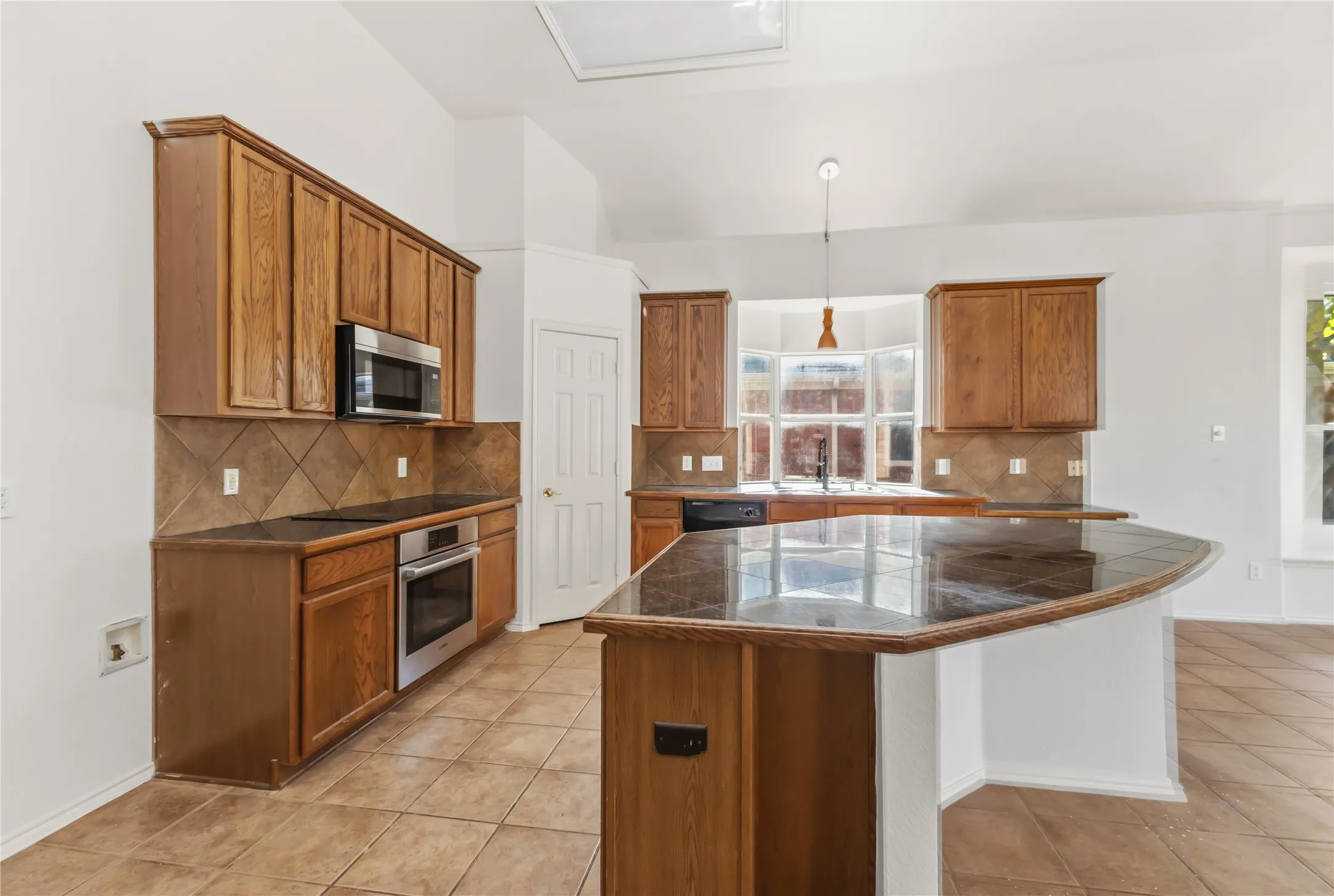 Kitchen featuring wood cabinetry, stainless steel appliances, dark countertops, tiled backsplash, and a central island with an integrated breakfast bar