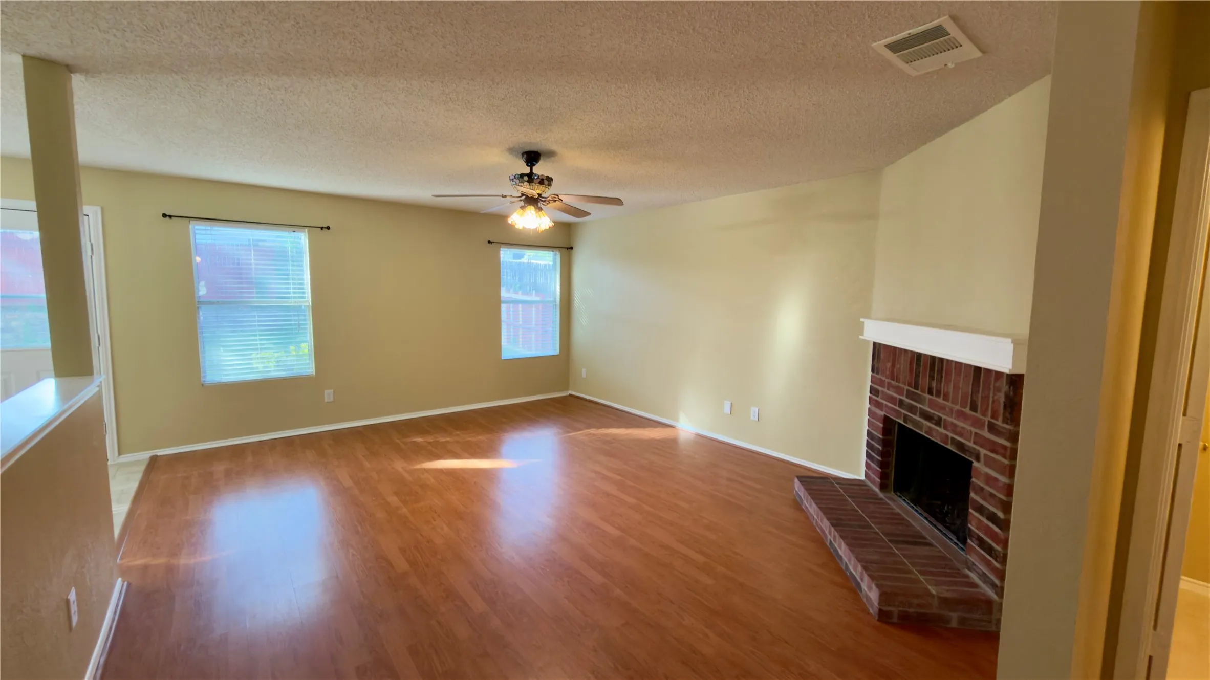 Spacious living area featuring wood-finish flooring, a brick fireplace with a white mantel, ceiling fan, and multiple windows with blinds