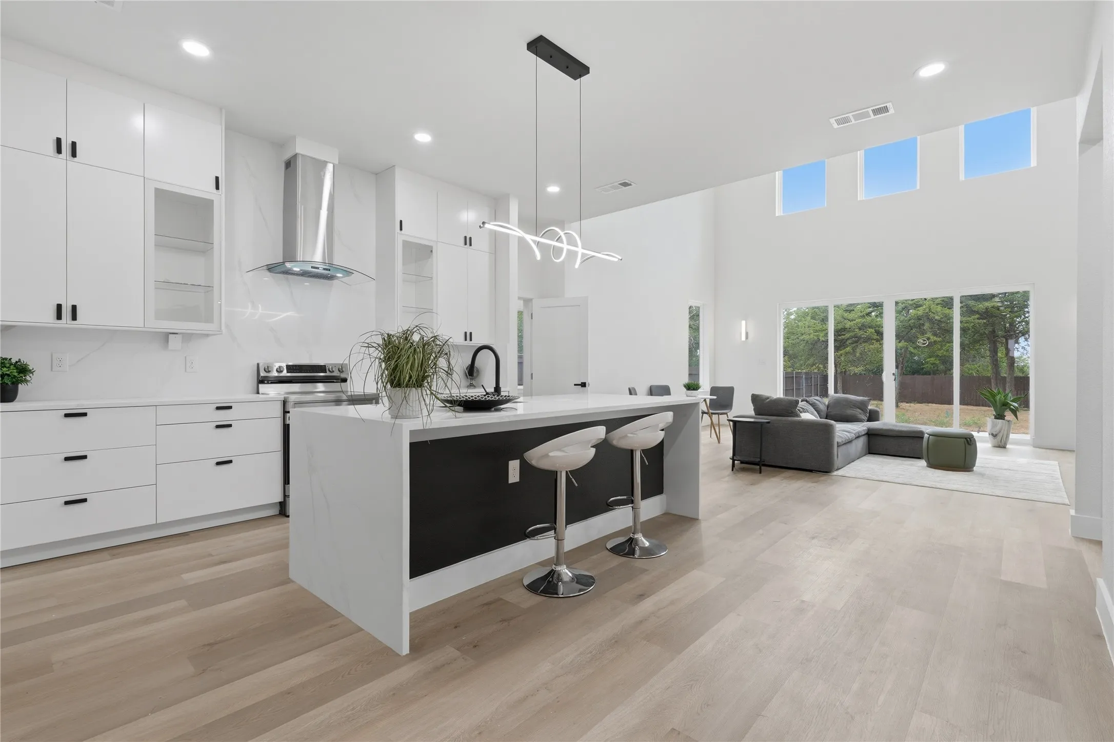 Open-concept layout featuring wood-finish flooring, a white kitchen with a waterfall island, stainless steel appliances, and a two-story living area with high windows and glass doors