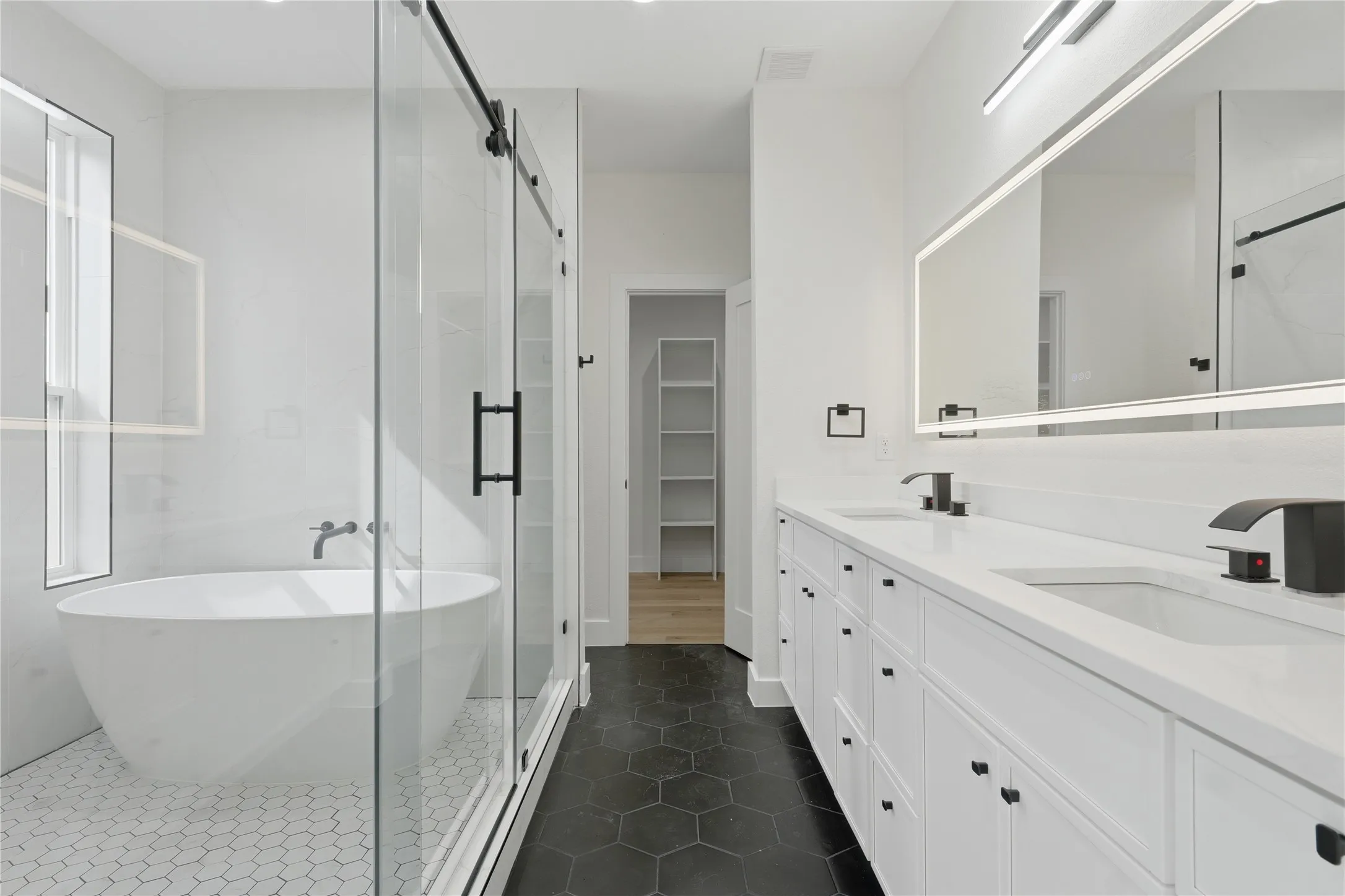 Freestanding soaking tub, glass-enclosed shower with sliding barn door, dual vanity with undermount sinks, illuminated mirror, and black hexagonal floor tiling
