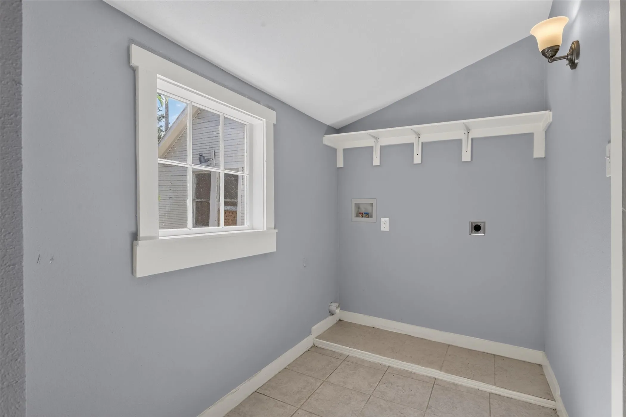 Utility area featuring a window with white trim, built-in shelving, a wall-mounted light fixture, tile flooring, and light gray walls