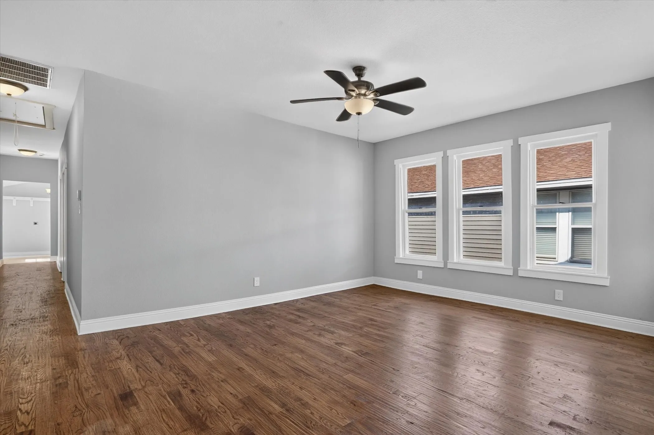 Spacious room featuring rich wood-finish flooring, three large windows with white trim, a ceiling fan with integrated lighting, and light gray wall finishes
