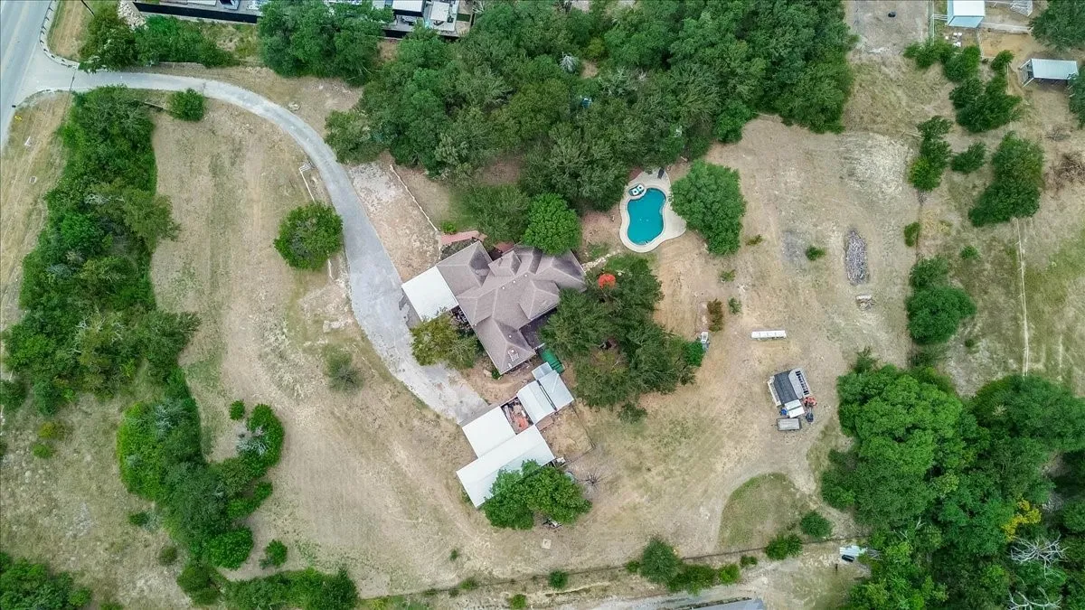 Expansive property featuring a main residence with a multi-gabled roof, an irregular-shaped swimming pool, extensive natural landscaping, a long driveway, and additional outbuildings