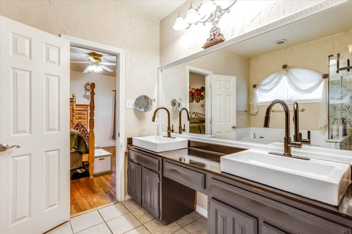Dual vessel sinks with dark bronze fixtures, dark wood-finish vanity, large vanity mirror, and neutral tile flooring