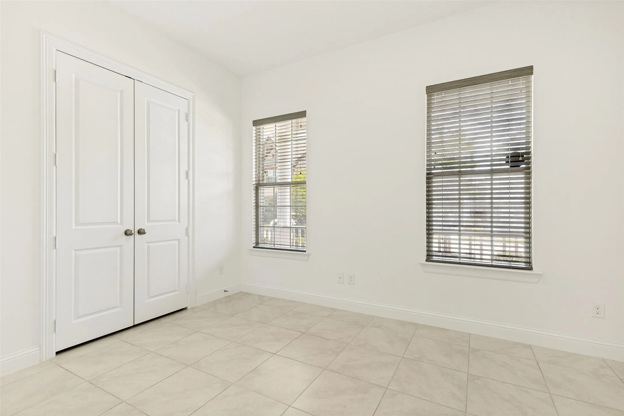 Bright interior space featuring light-toned tile flooring, two windows with horizontal blinds, and white bi-fold doors