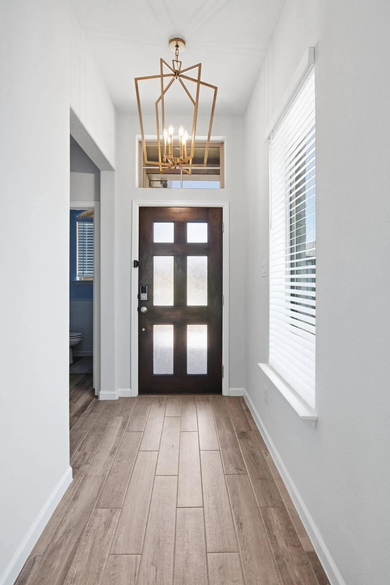 Entryway featuring wood-finish flooring, a contemporary geometric chandelier, a multi-paneled door with frosted glass inserts, a transom window, and a window with horizontal blinds
