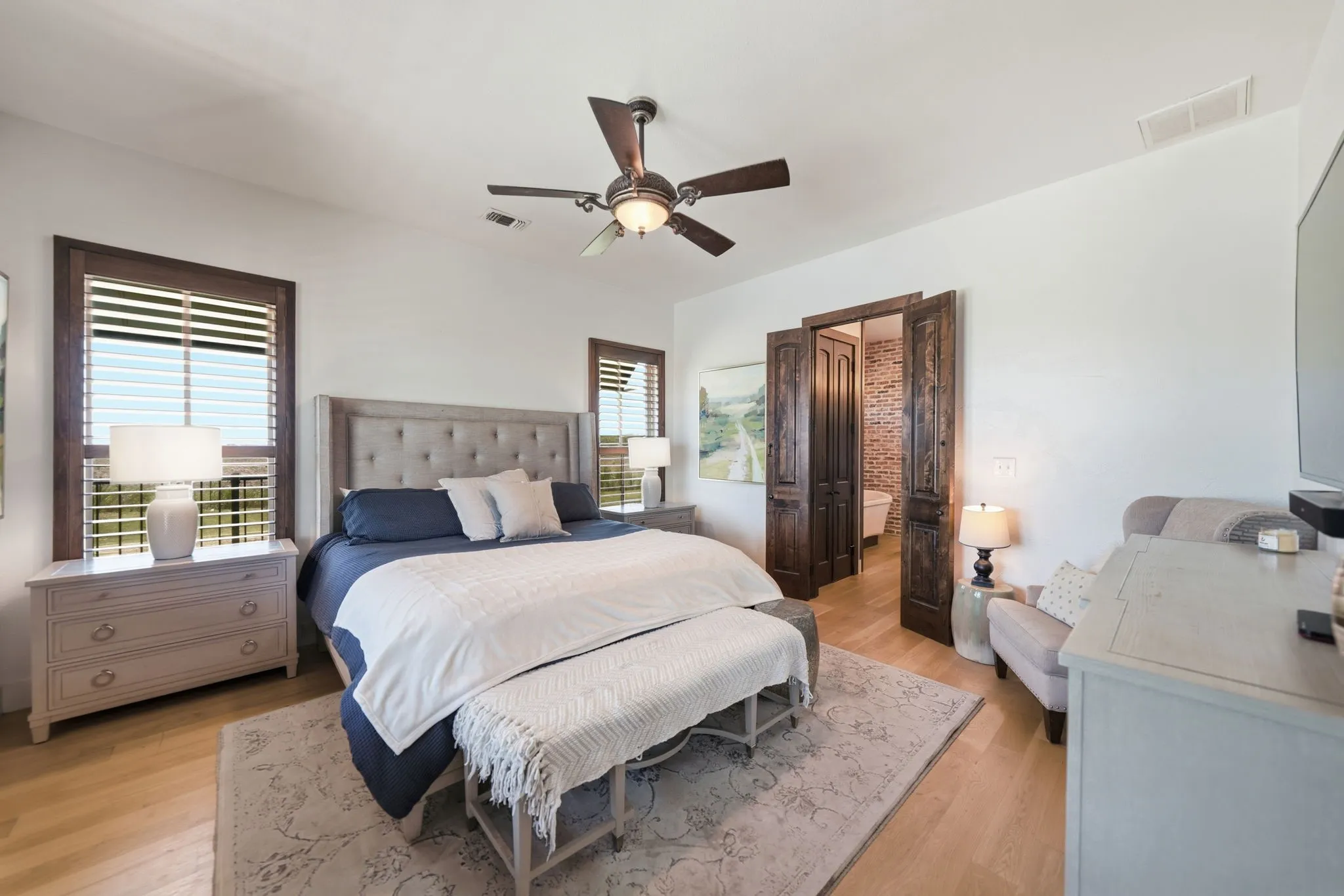 Spacious primary bedroom featuring light wood-finish flooring, a ceiling fan with light fixture, and two windows with wood-framed trim and blinds