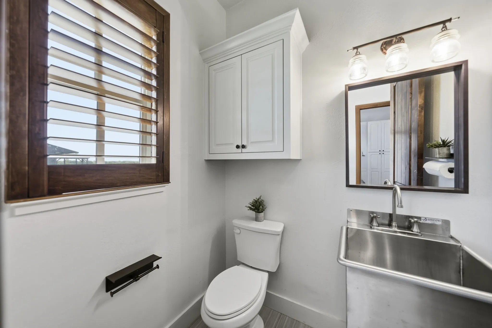 Bathroom featuring a dark wood framed window with horizontal blinds, white wall cabinetry, a white toilet, and a stainless steel utility sink with a wall-mounted faucet