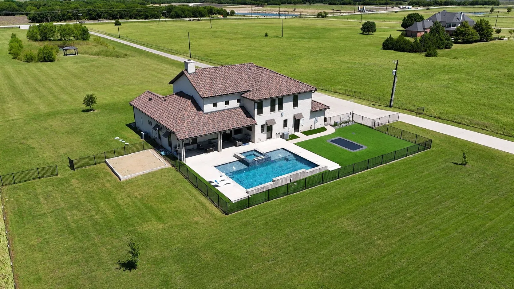 Expansive property featuring a contemporary home with a red tile roof, an in-ground swimming pool, a separate spa, a fenced lawn area with a trampoline, and a sand court