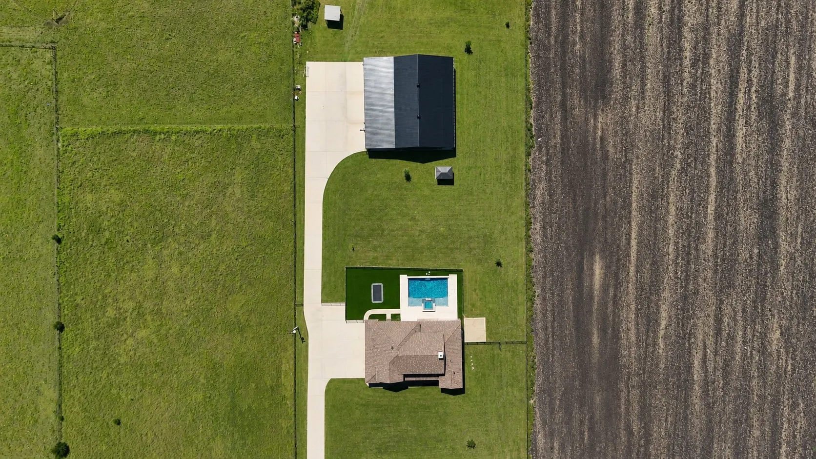 Expansive property featuring a main residence with a brown shingle roof, a detached black-roofed structure, a swimming pool with surrounding patio, a concrete driveway, and extensive green space