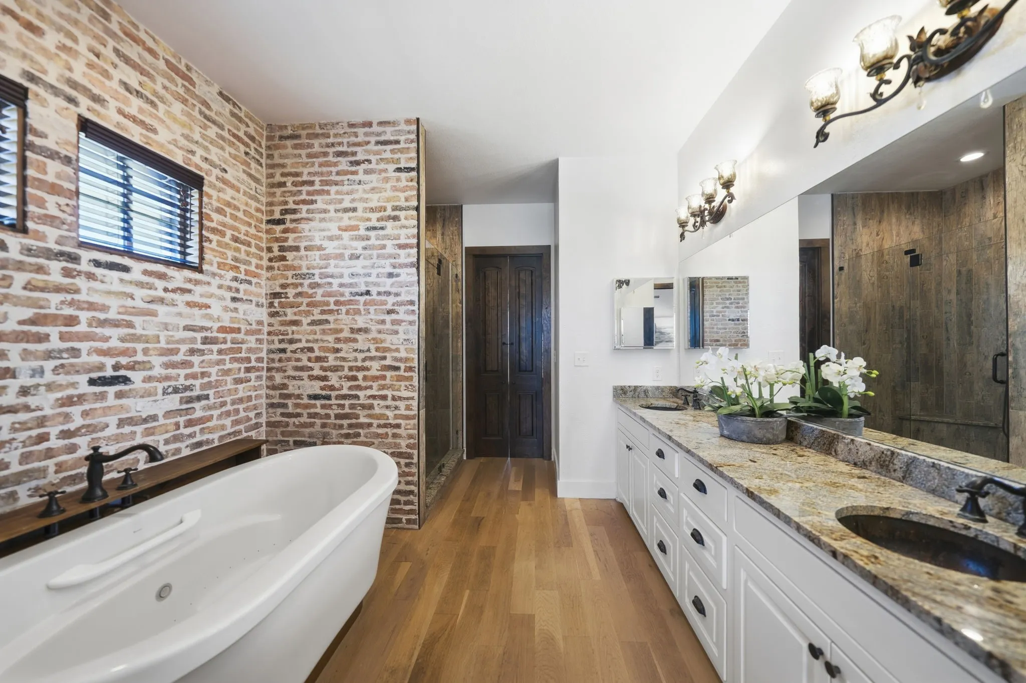 Bathroom featuring a freestanding soaking tub with wall-mounted faucet, an exposed brick accent wall, wood-finish flooring, a double vanity with stone countertop and integrated sinks, and a glass-enclosed shower with stone-look wall tiles