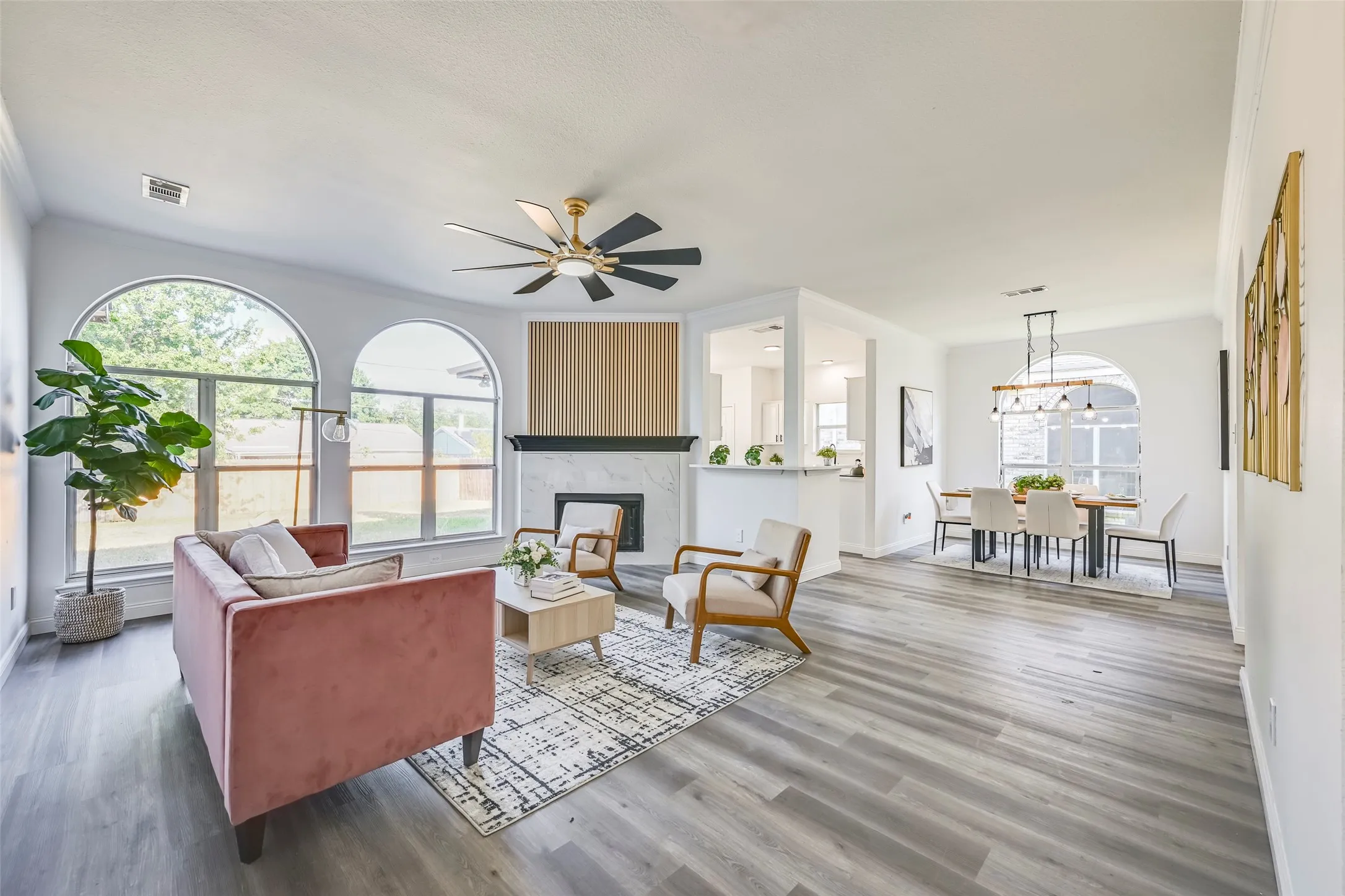 Expansive living area with wood-finish flooring, featuring a fireplace with a white stone surround, a wood-paneled accent wall, and a contemporary ceiling fan
