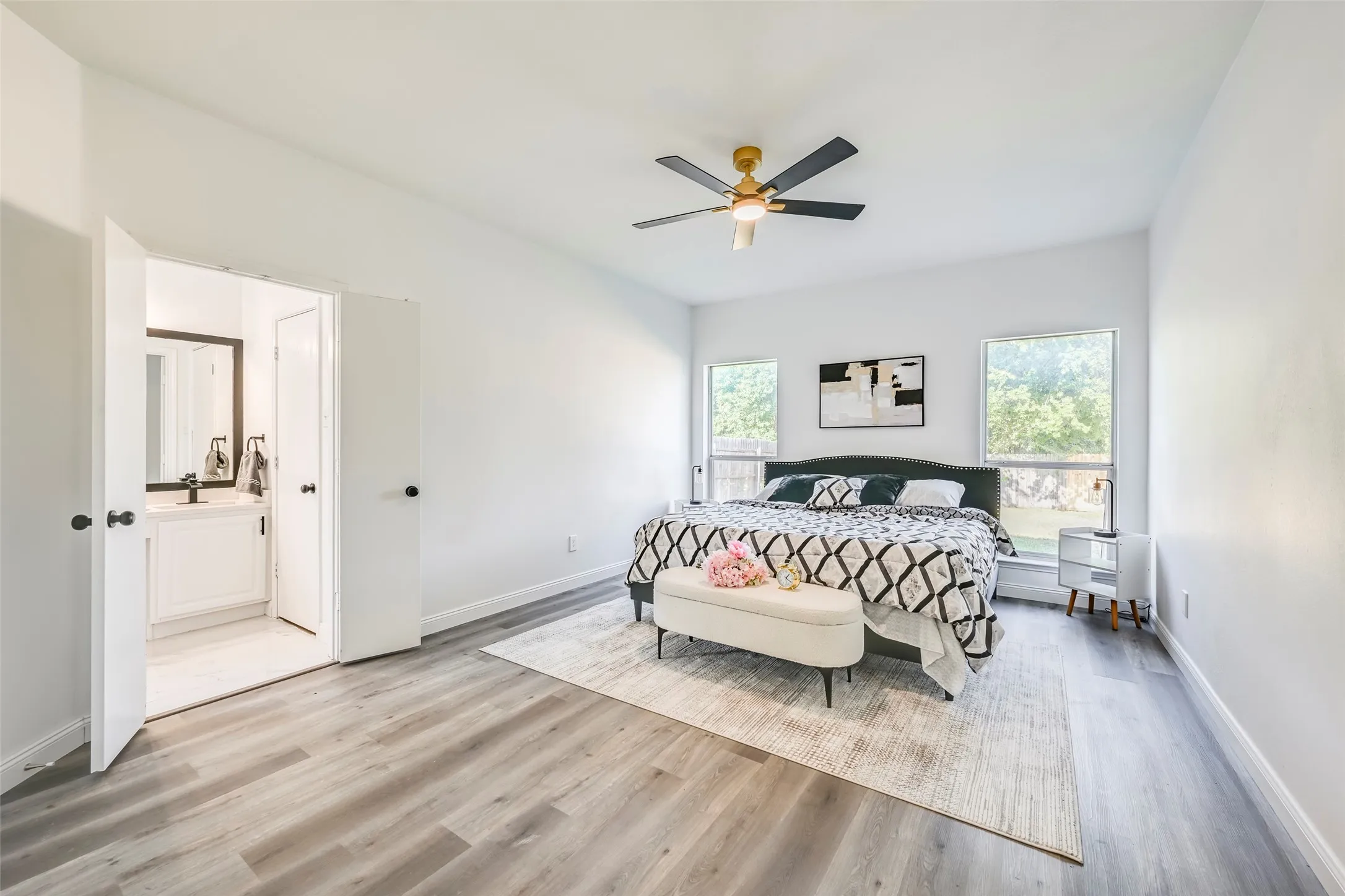 Spacious bedroom featuring light wood-finish flooring, a ceiling fan with integrated lighting, and two large windows
