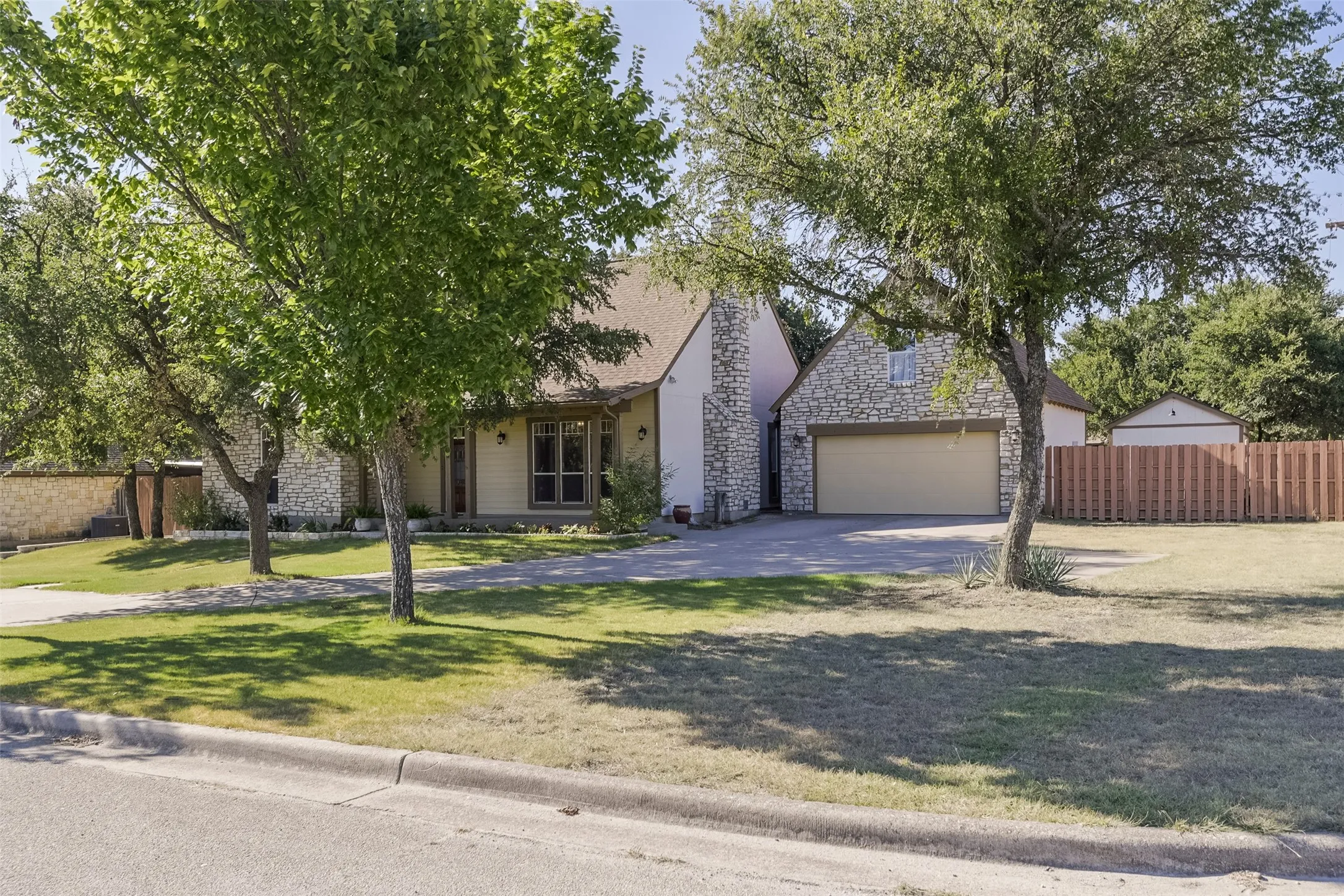 Stone veneer exterior with a prominent two-car garage, multiple mature trees, a manicured lawn, and a wooden privacy fence
