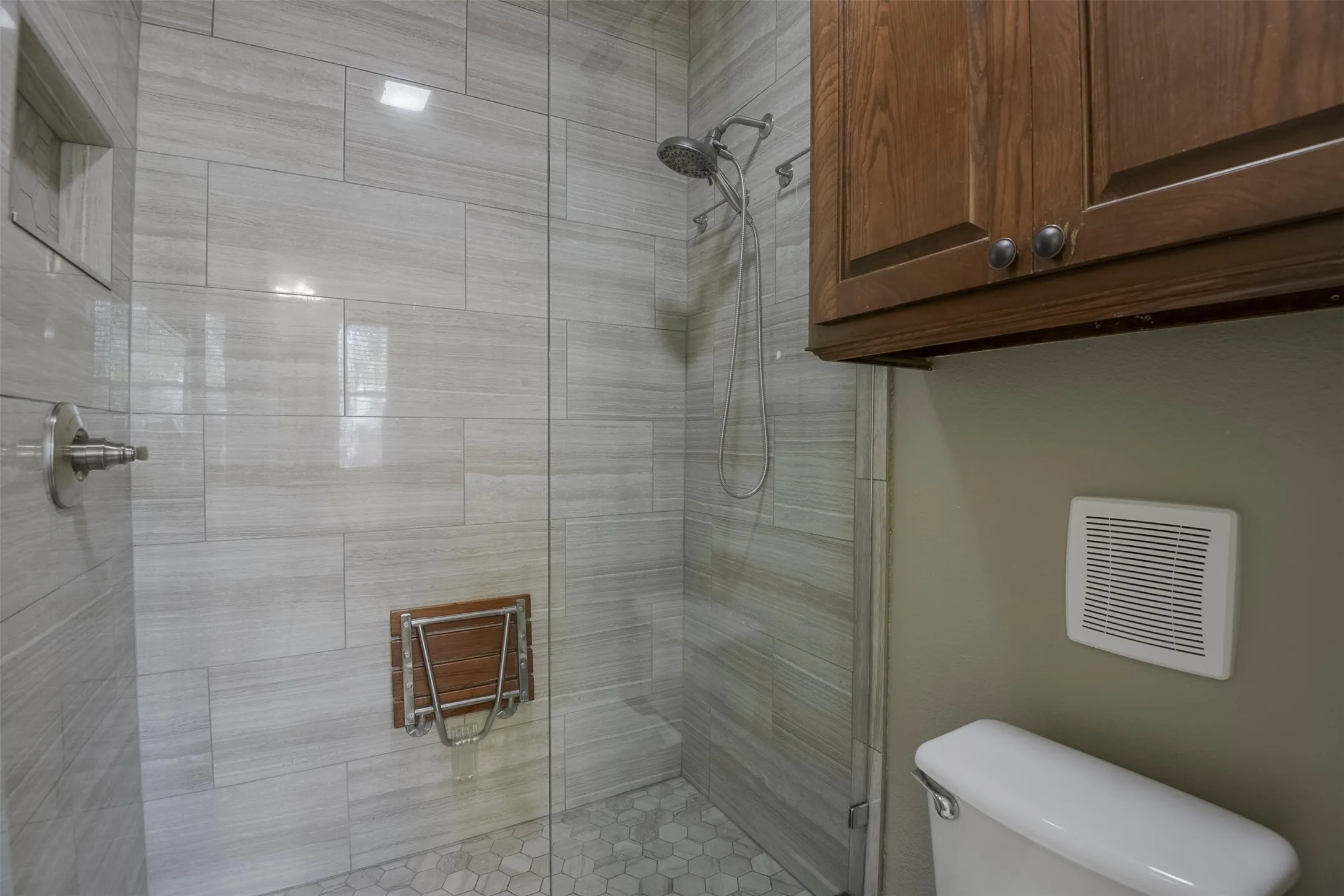 Glass-enclosed shower featuring light grey horizontal wall tiles, a built-in shower niche, a fold-down wood-finish shower seat, and a hexagonal tile floor