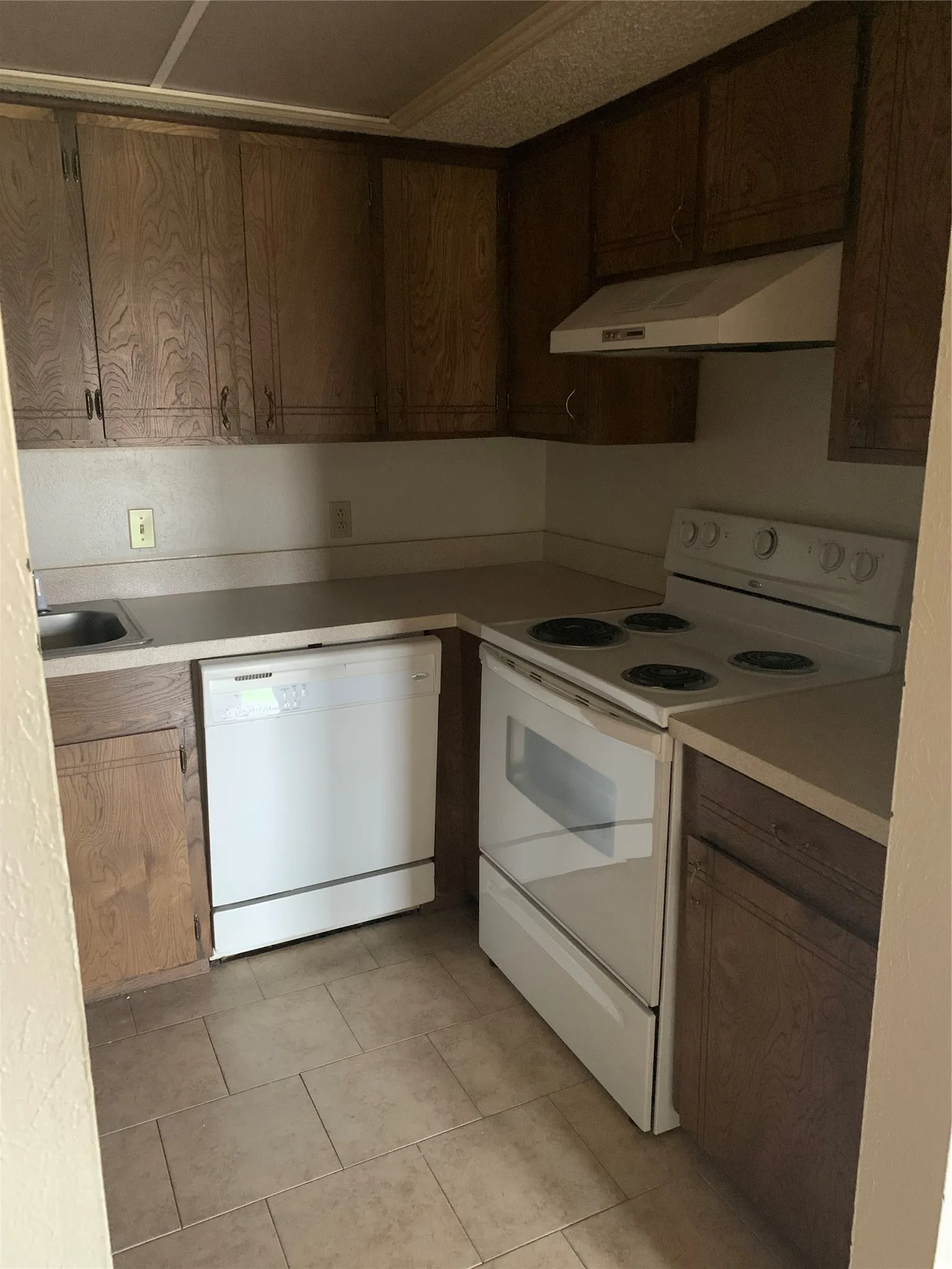 Kitchen featuring wood-finish cabinetry, light-toned countertops, tile flooring, a built-in dishwasher, and a white range with an overhead vent hood