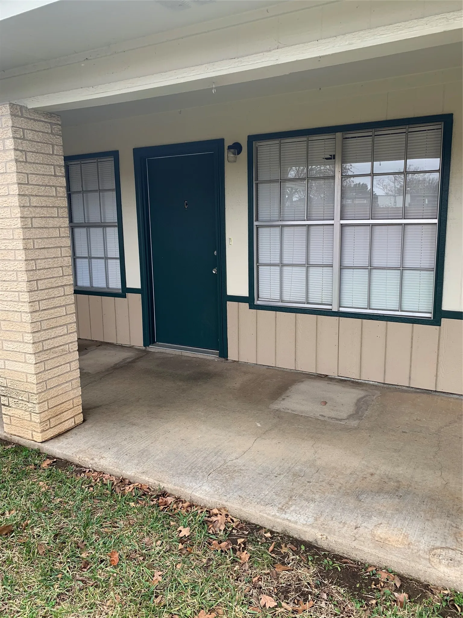 Covered concrete porch with a brick pillar, dark green entry door, and multi-pane windows