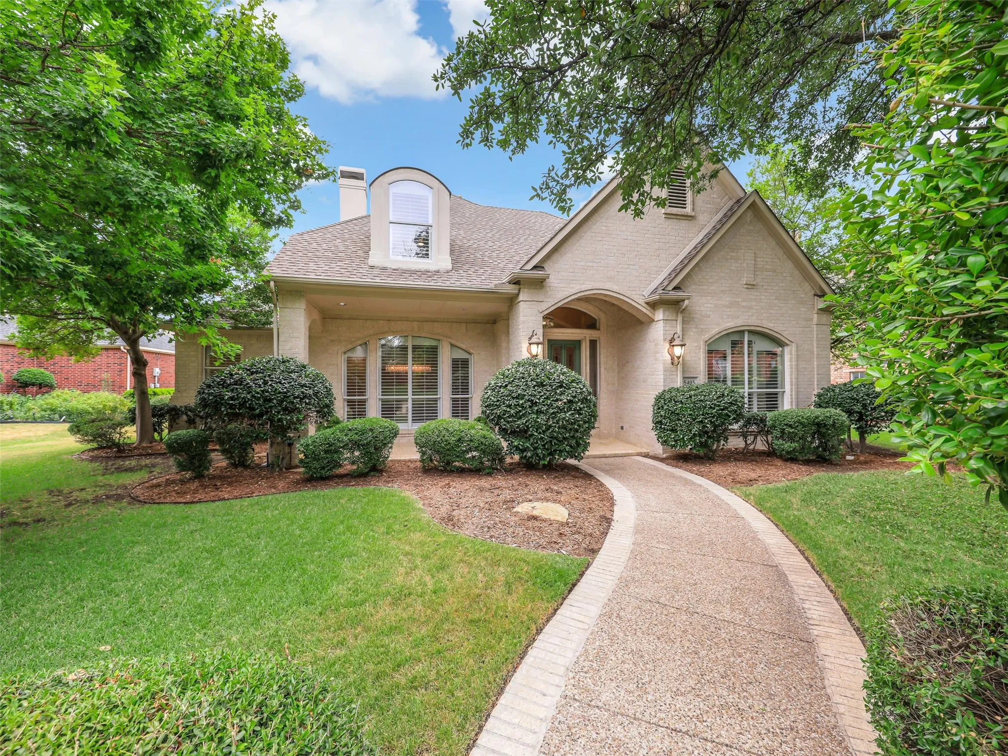 Cream-colored brick exterior with arched entryway, dormer window, and a curved exposed aggregate walkway with brick edging