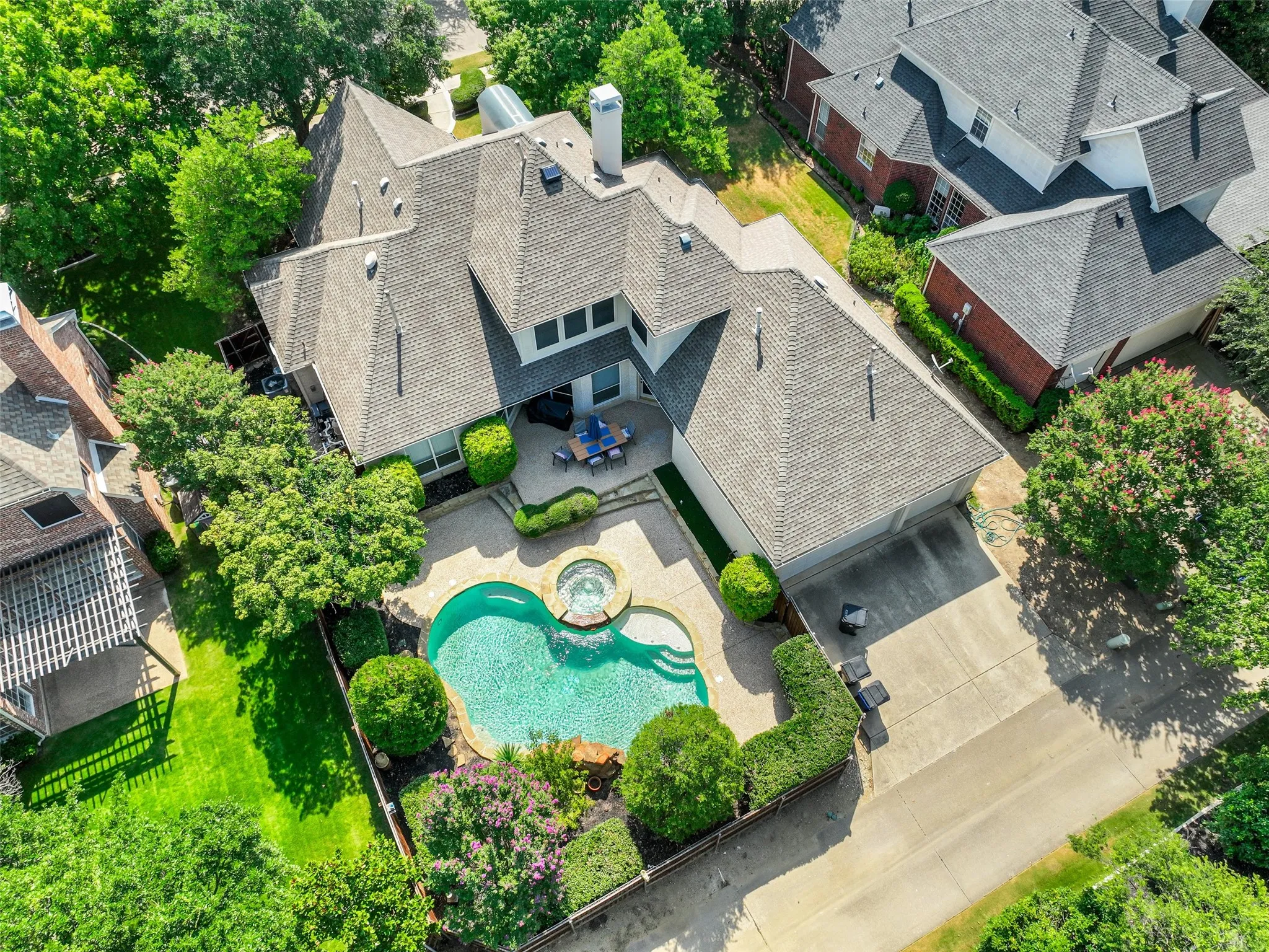 Aerial perspective showcasing a multi-level roofline, an irregular-shaped swimming pool with an integrated spa, a concrete patio, a concrete driveway, and mature landscaping