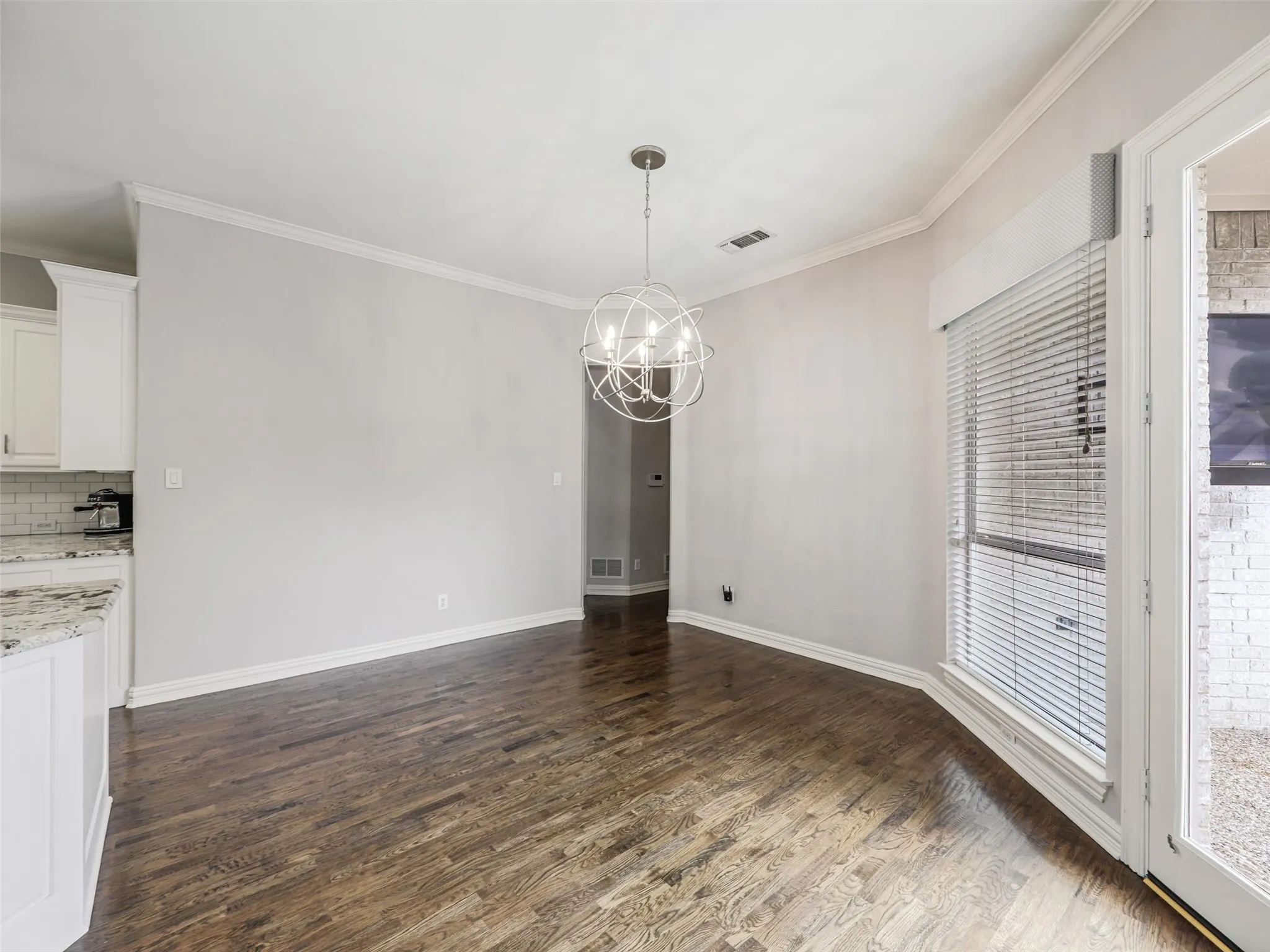 Wood-finish flooring, crown molding, modern spherical chandelier, white shaker cabinetry, and light-toned countertop with subway tile backsplash