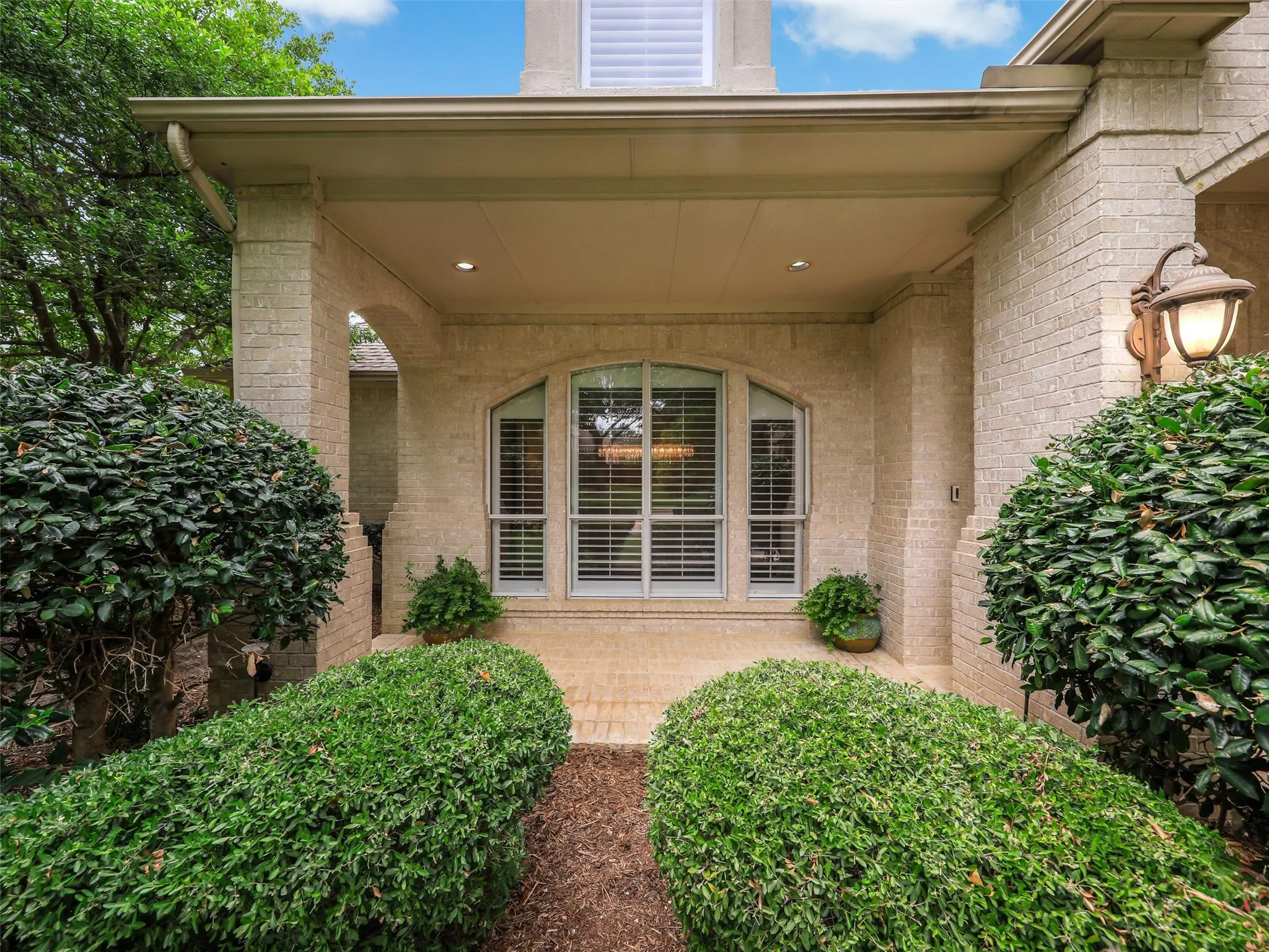 Covered entry with recessed lighting, light-colored brick exterior, arched window with horizontal blinds, and a brick paver pathway