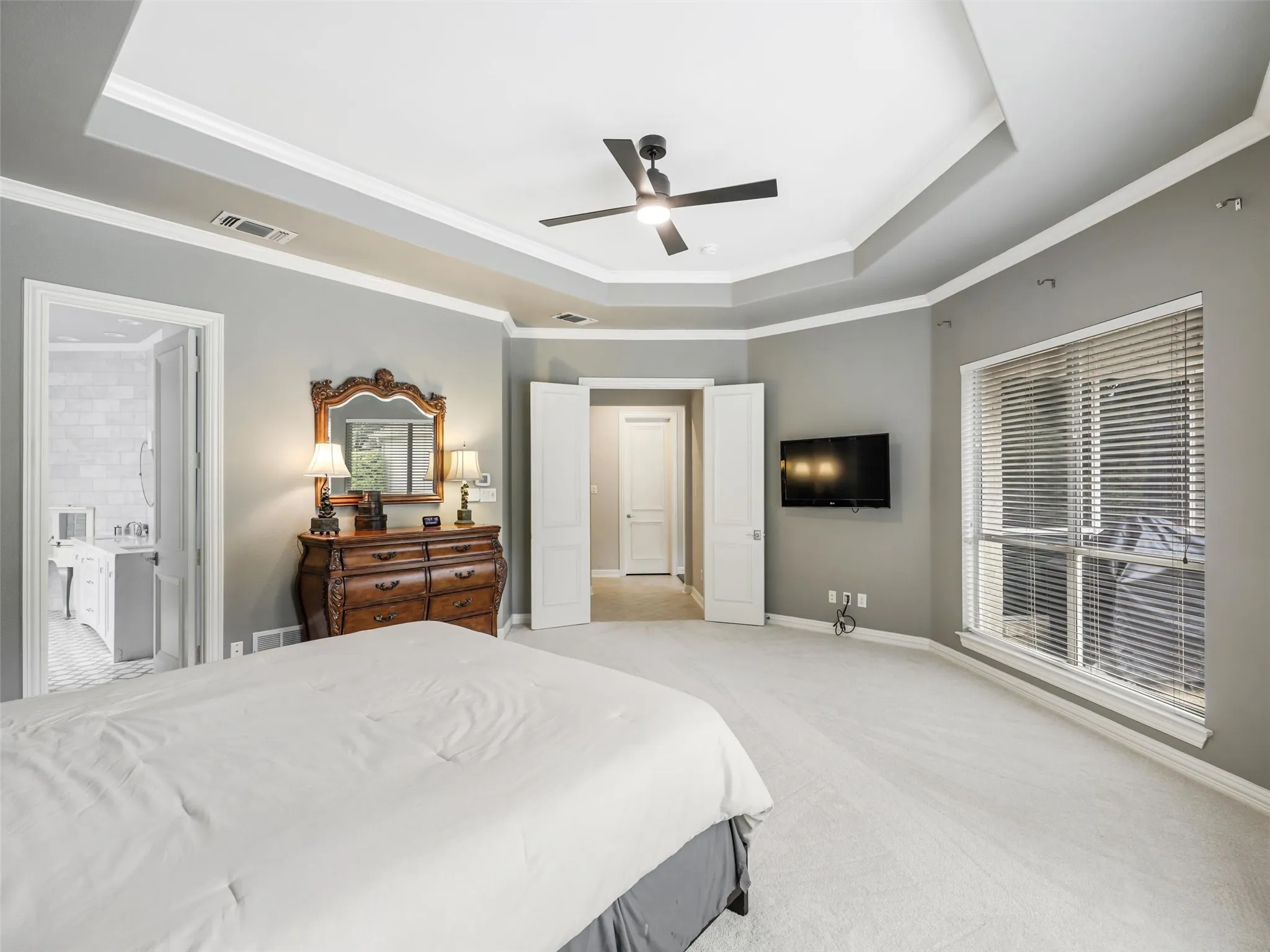 Spacious primary bedroom featuring a tray ceiling with ceiling fan, light-filled windows with plantation shutters, hardwood-finish flooring, and an ensuite bathroom