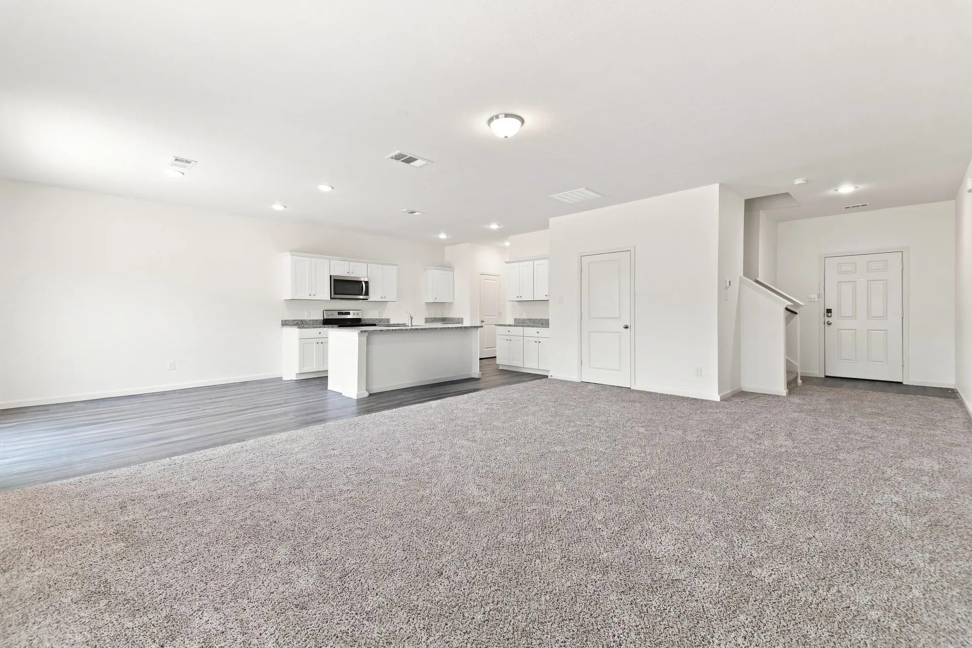 Spacious living area featuring light gray carpeting and a kitchen with white cabinetry, stainless steel microwave, and an island with a countertop