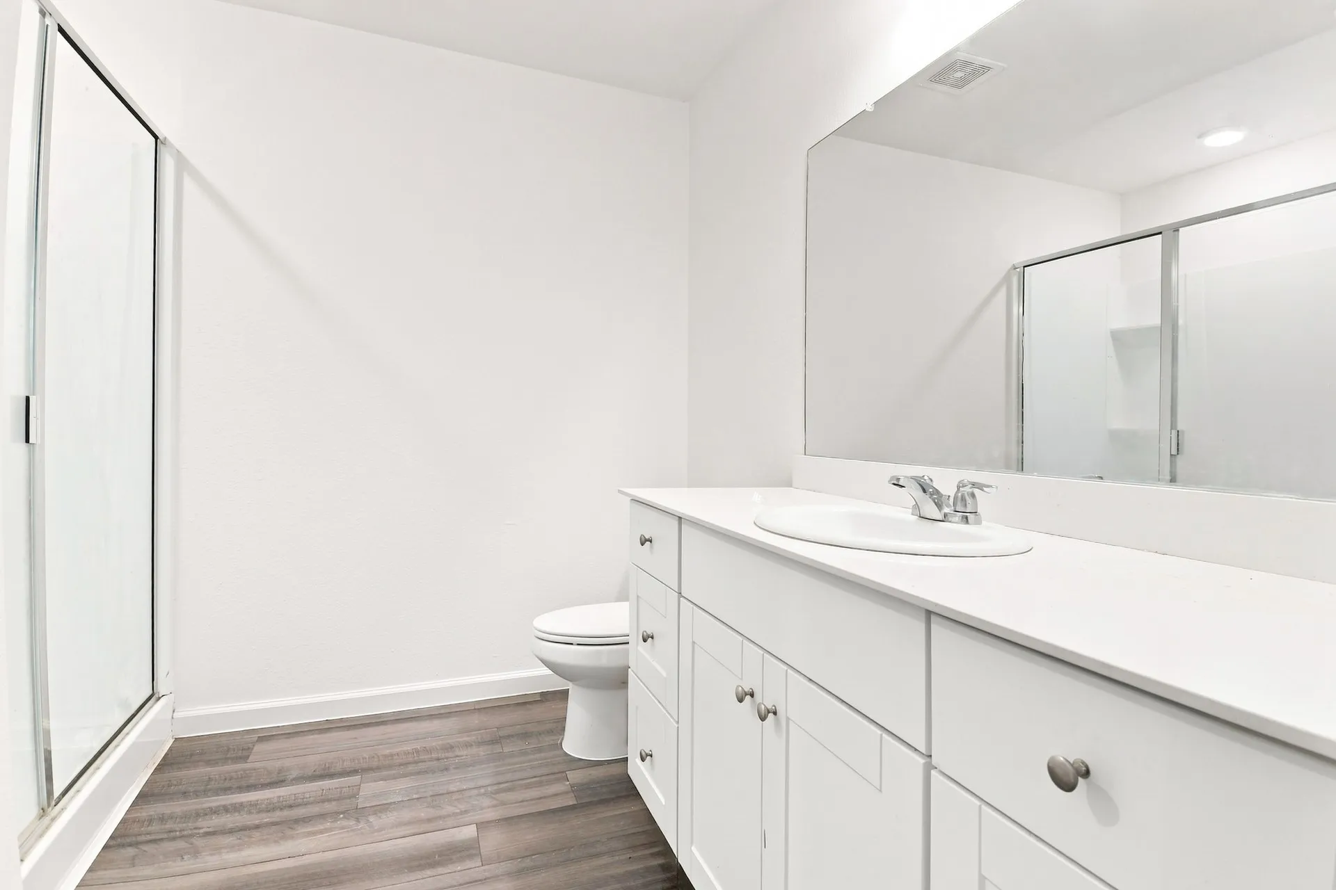 Bathroom featuring a spacious vanity with white cabinetry and an integrated sink, a large wall-mounted mirror, a glass-enclosed shower, and wood-finish flooring